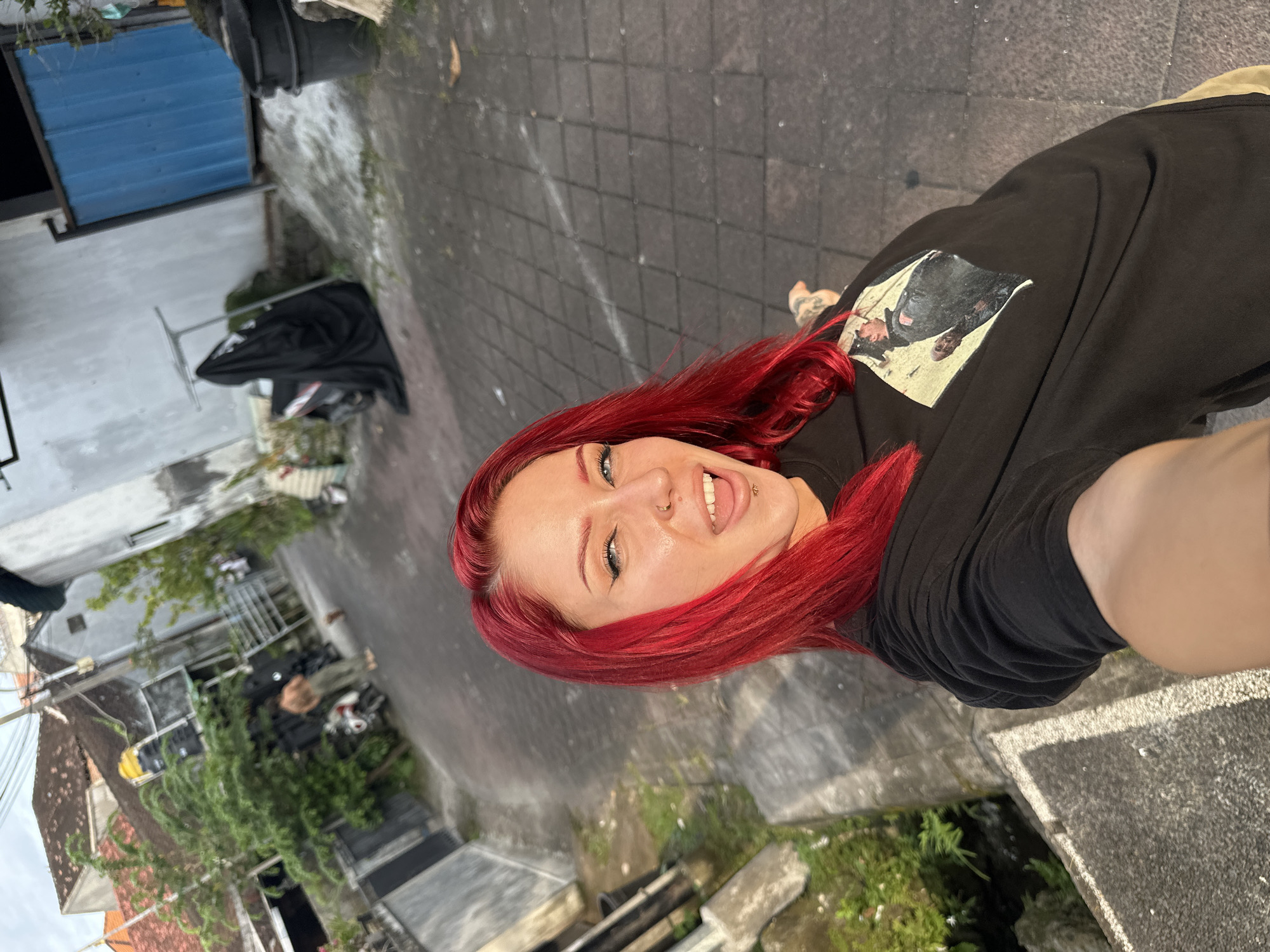 A woman with bright red hair, blue eyes, black eyeliner, a nose piercing, and a labret piercing is depicted in a selfie, looking directly at the camera with her mouth open and tongue slightly extended. She wears a black t-shirt featuring a light-colored graphic print of a dark-clad figure, and a tattoo is visible on her right upper arm.
The scene is set in a paved alleyway or residential street within Kerobokan, Indonesia. The ground is covered with grey interlocking pavers. To the left, a narrow concrete ditch contains green vegetation, including ferns and moss. Buildings line both sides of the street, displaying light-colored walls, some with visible terracotta roof tiles. A building on the right features a prominent blue corrugated metal gate or door.
In the mid-ground, a shirtless male in light-colored pants stands near a white and red scooter parked on the street. Another scooter is partially covered by a black sheet next to a metal clothes drying rack. Black refuse bins are visible near structures. Sparse trees and bushes are present. The lighting is natural, consistent with daytime.