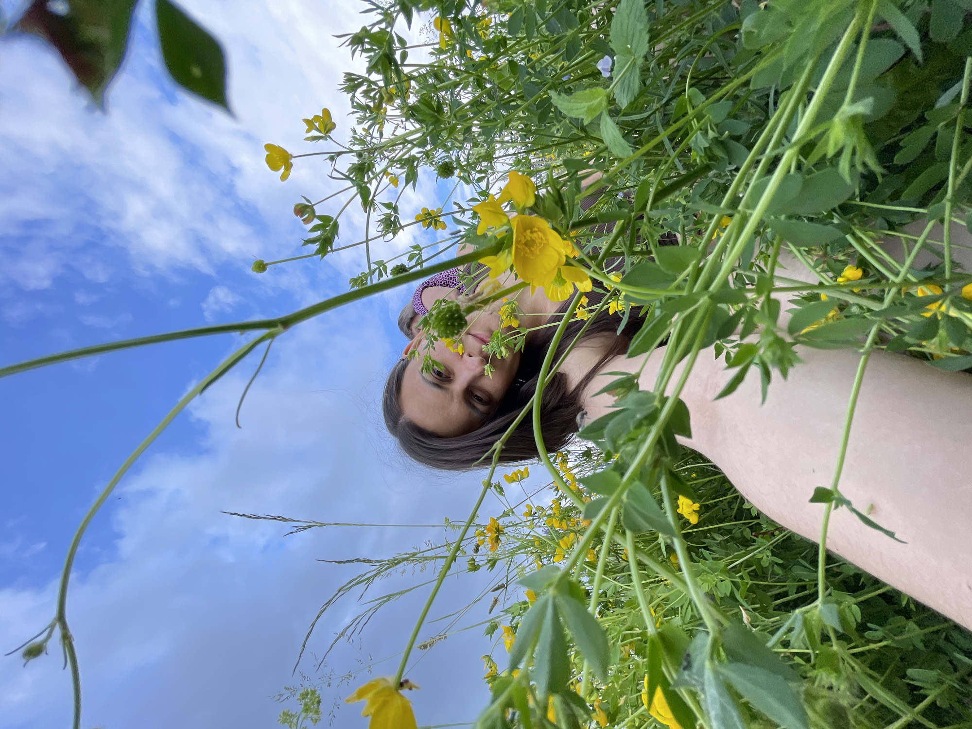 A low-angle, wide shot captures a person lying in a field of yellow wildflowers and green grass under a cloudy blue sky. The person's arm and shoulder are visible in the foreground, with their face partially obscured by the foliage as they look towards the camera. The background is dominated by the sky and the surrounding greenery. The location is identified as Primorsky Krai, Russia.