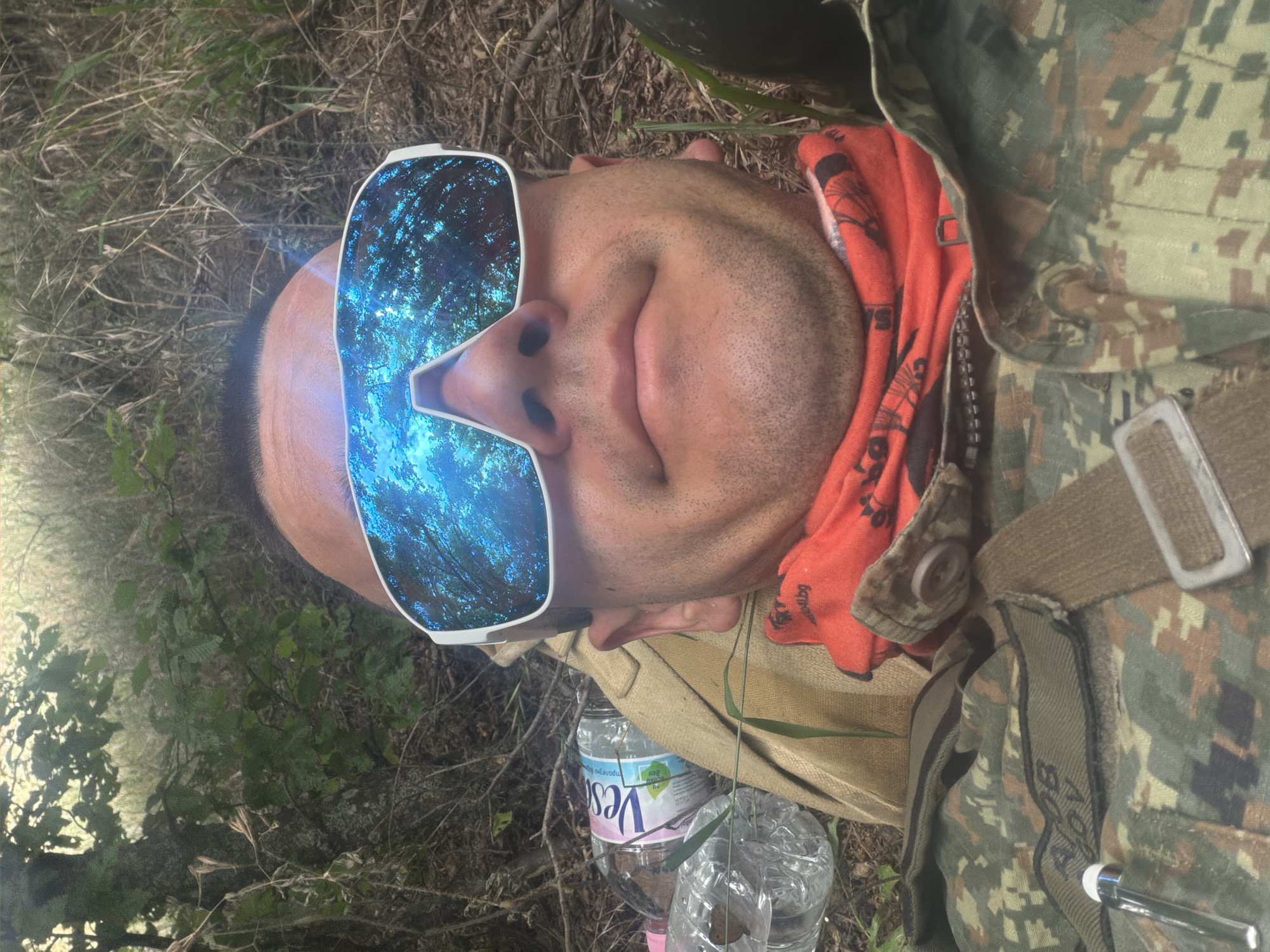 This media file is a close-up, low-angle shot of a person lying on the ground in a wooded or grassy area, possibly Beliakovets, Bulgaria. The person is wearing sunglasses with a reflective lens that shows a blue, starry sky or a forest canopy. They are also wearing a camouflage uniform, an orange bandana around their neck, and a belt. Two plastic water bottles are visible next to them. The lighting suggests it is daytime, and the environment appears natural and somewhat wild. The person's face is relaxed, and they seem to be at rest. The camouflage pattern and the presence of the water bottles might indicate an outdoor activity such as hiking, camping, or military training. The overall impression is one of a person taking a break in a natural setting. Visible text on the orange bandana appears to be "BOG" and "BLOG". The label on one of the water bottles is partially visible and reads "Ves".