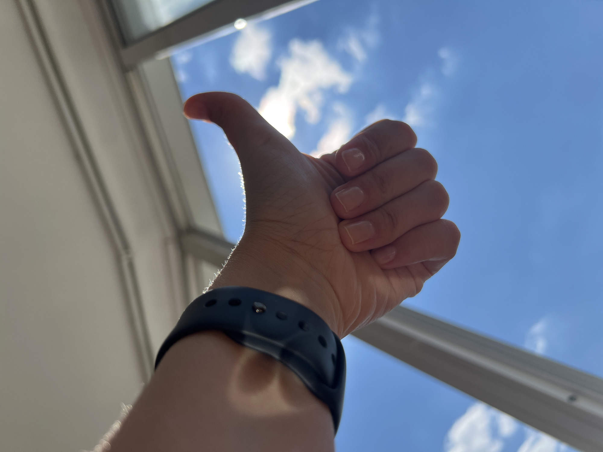 A person's hand is held up with the thumb extended in a "thumbs up" gesture. A dark blue wristband is visible on the person's wrist. The background is a bright blue sky with scattered white clouds, suggesting an outdoor scene or a view from a window. The image is backlit, creating a silhouette effect on the hand and arm. There is no discernible location in Tolyatti, Russia, or any other city.