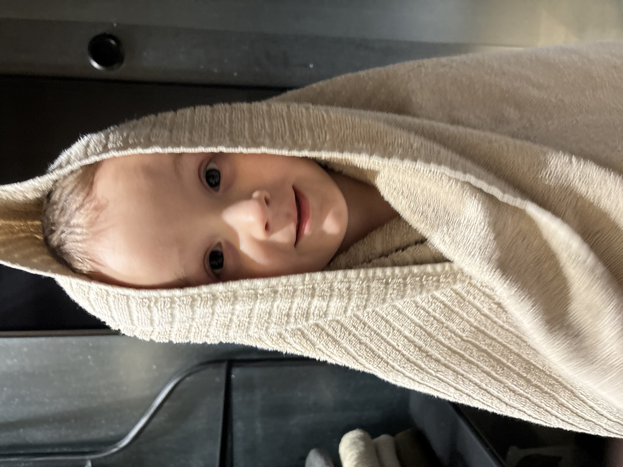 A young child, appearing to be a toddler, with fair skin, light hair, and dark eyes, is depicted looking directly forward with a slight smile. The child is wrapped from head to lower body in a light beige, textured terrycloth towel, which forms a hood. The towel exhibits a subtle ribbed pattern. In the background, to the left of the child, a stack of folded towels in various shades of light grey and beige rests on a dark, flat surface. Behind this, a dark, vertical panel shows a dark, curved cable or pipe. To the right, another dark vertical surface features a circular dark fitting. The indoor environment appears to be a functional space, possibly a bathroom or changing area, located in Chaikovsky, Russia.
