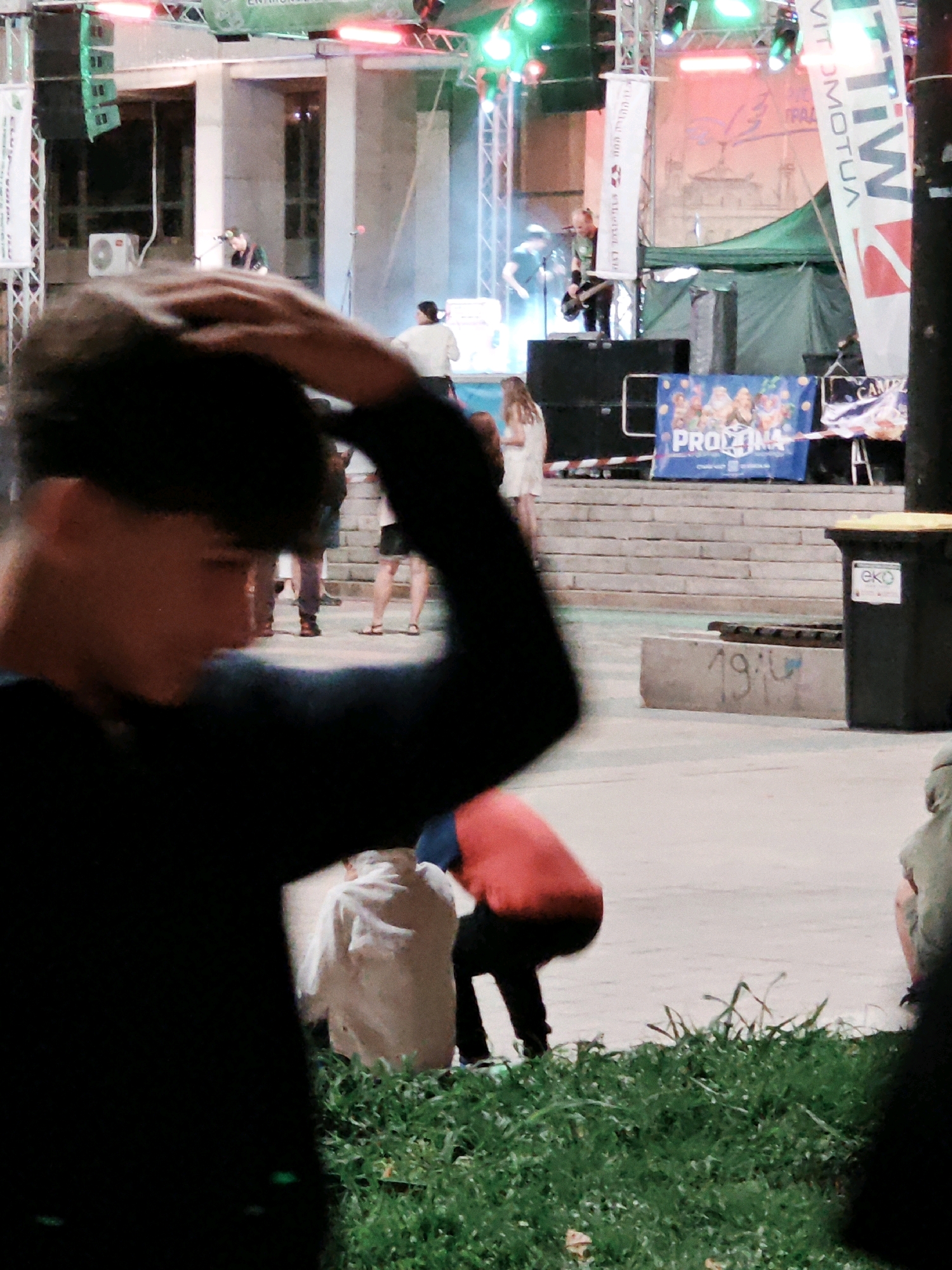 In an outdoor urban setting, a blurred foreground features a person in a black shirt with their hand raised to their head. Behind them, a stage is set up with lights and speakers. On the stage, a musician in a green shirt is playing a guitar, and another person is standing to their left. A banner with colorful images and text reading "PROIZINA" is displayed in front of the stage. To the right, a black trash can with a yellow lid is visible, and in the background, there are steps leading up to a building. Several other people are present, some blurred and out of focus, appearing to be spectators. The ground is paved, and there is a patch of green grass in the foreground. The lighting suggests it is nighttime or dusk, with stage lights providing illumination.