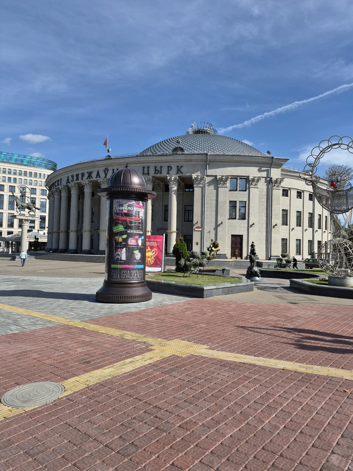 The image captures a daytime view of the Belarusian State Circus in Minsk, Belarus. The grand, neoclassical building features a prominent dome and a colonnade lining its facade. In the foreground, a tall, cylindrical advertisement pillar displays various graphics and text, including an image of a red car. The area in front of the circus is paved with reddish-brown bricks and features yellow tactile paving. To the left, a statue of a horse and rider stands on a pedestal, and a person is walking away from the camera. To the right, a whimsical metal sculpture resembling a clown or jester is visible. The sky is blue with scattered clouds and a contrail. The scene appears to be a public square in front of a significant cultural landmark.