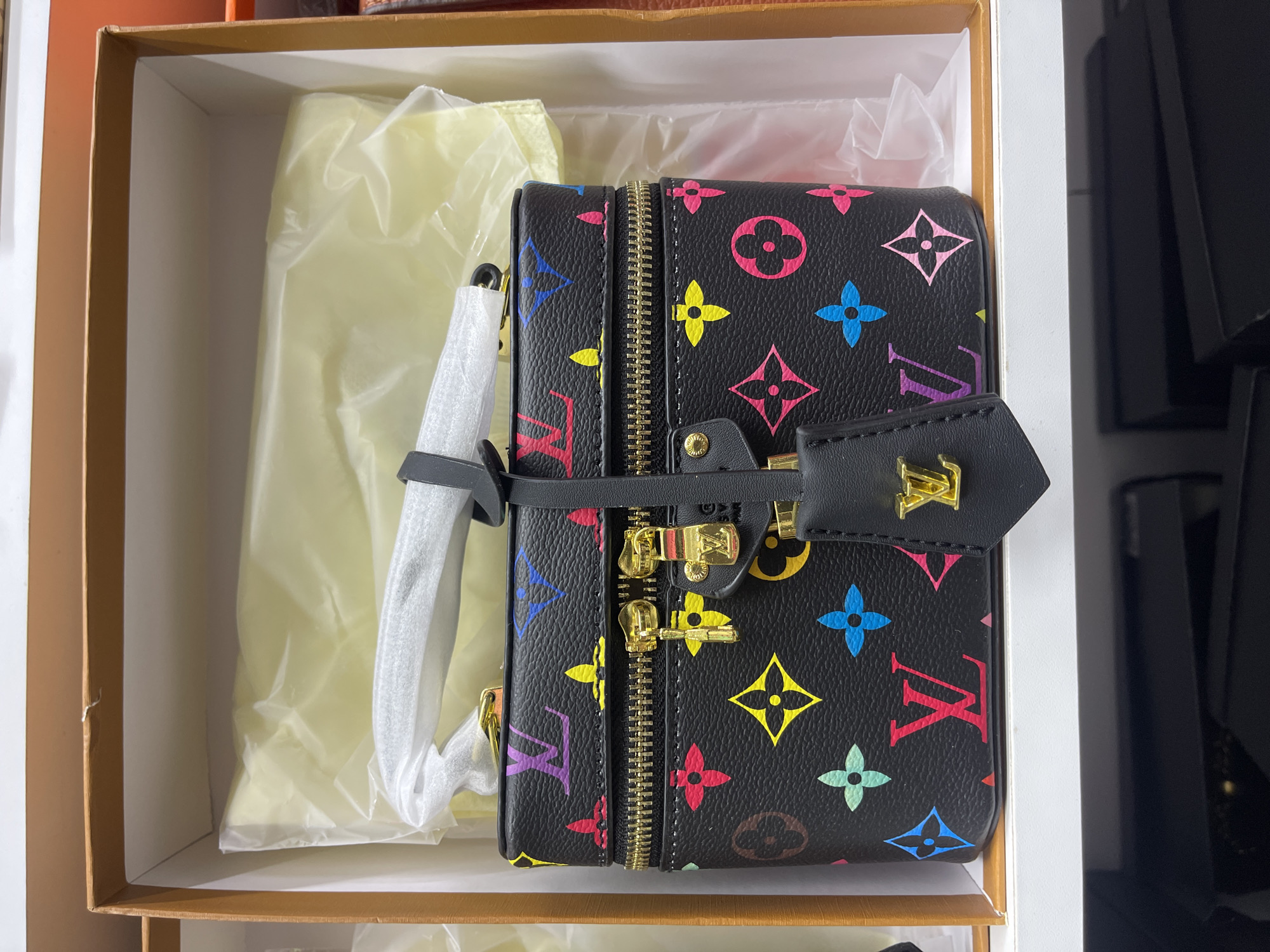 A black Louis Vuitton vanity case with the brand's multicolored monogram pattern is displayed inside a cardboard box. The case has a black strap attached to its zipper pull, which also features a gold-toned LV emblem. The box is open, and the case is nestled within light-colored tissue paper. The background suggests an indoor setting, possibly a retail environment, with shelving visible below. No people are present in the image. The location of Moscow, Russia, is not determinable from the visual information provided.