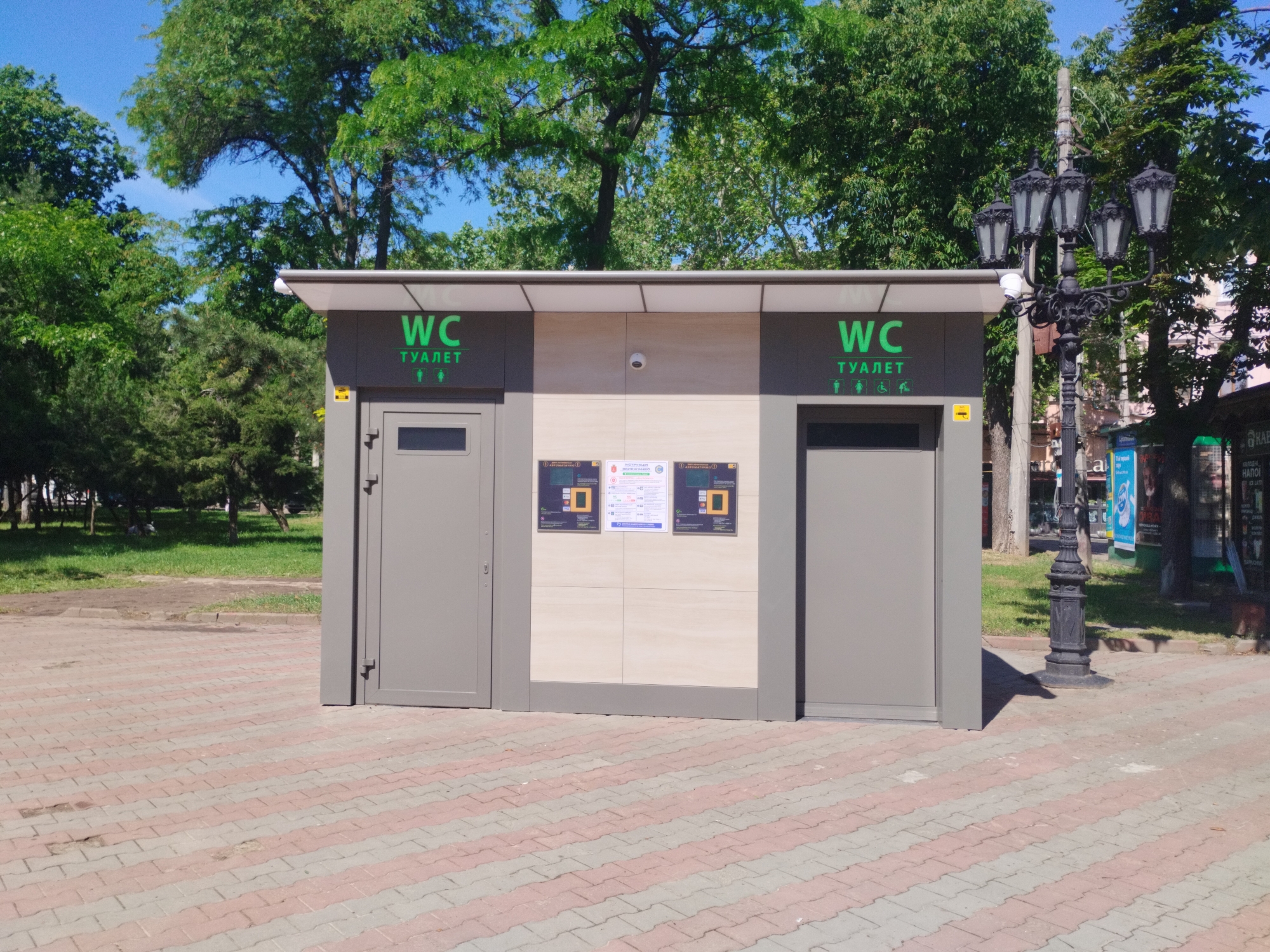 A public toilet stands on a brick-paved area with trees and greenery in the background. The toilet structure is gray with a lighter beige section in the middle, flanked by two gray doors. Each door has the letters "WC" and the word "туалет" (toilet in Ukrainian) printed in green above it, accompanied by symbols representing male, female, and disabled users. A lamp post with five ornate lanterns stands to the right of the toilet. The location is Odesa, Ukraine.