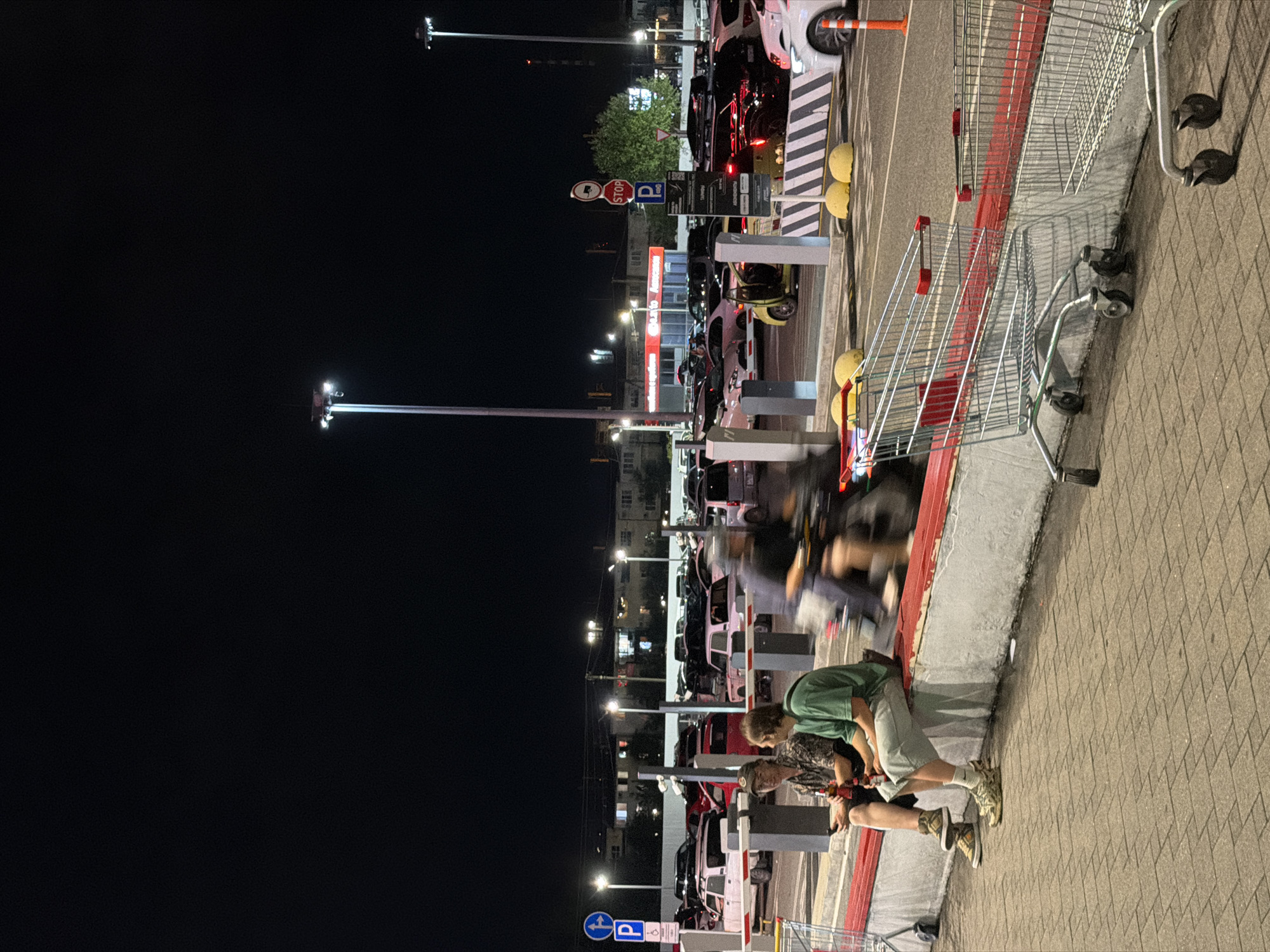 The image depicts a nighttime scene in a parking lot in Sirius, Russia. Two young men are seated on a low concrete barrier, one holding a bottle. In the background, a busy parking lot is visible with numerous cars, many of which appear to be parked. A motorcycle with a rider is captured in motion, creating a blur. Several shopping carts are present in the foreground, some of which are also in motion, indicating movement within the frame. Streetlights illuminate the area, casting a dim glow on the surroundings. The overall environment suggests a commercial or retail area with a parking facility.