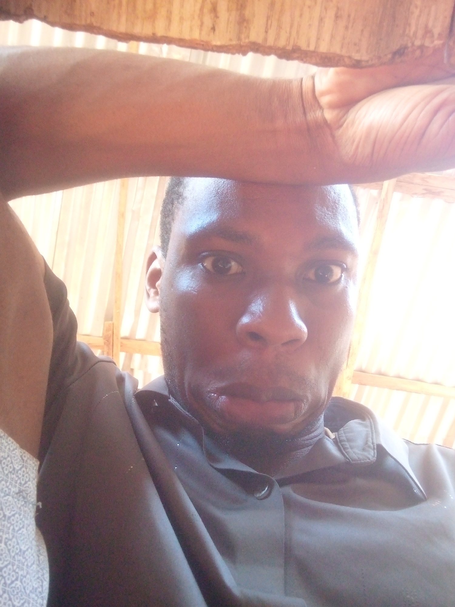A dark-skinned man is looking directly at the camera. His right arm is raised over his head, resting on a wooden structure. He is wearing a dark shirt with a buttoned collar. The background appears to be a brightly lit room with wooden beams. The location is Unknown, Unknown.