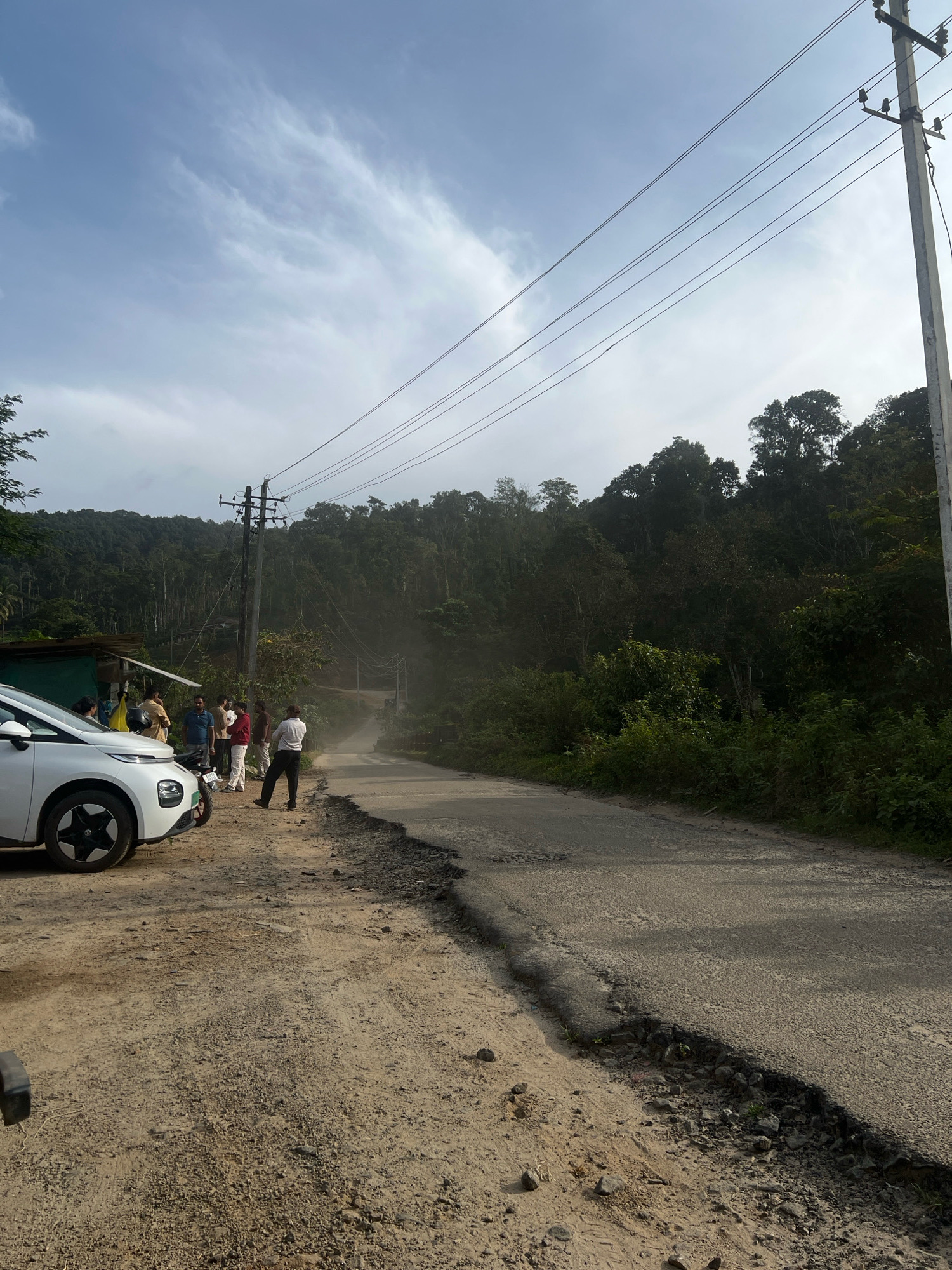 A group of people are standing on the side of a damaged asphalt road in a rural, forested area. A white car is parked on the dirt shoulder to the left of the road. A utility pole with power lines runs along the right side of the road. The road appears to be partially eroded, with visible damage to the asphalt and embankment. The surrounding environment is characterized by dense green foliage and trees on a hillside. The sky is partly cloudy, with a dusty haze visible in the distance down the road. It is daytime, with natural lighting.