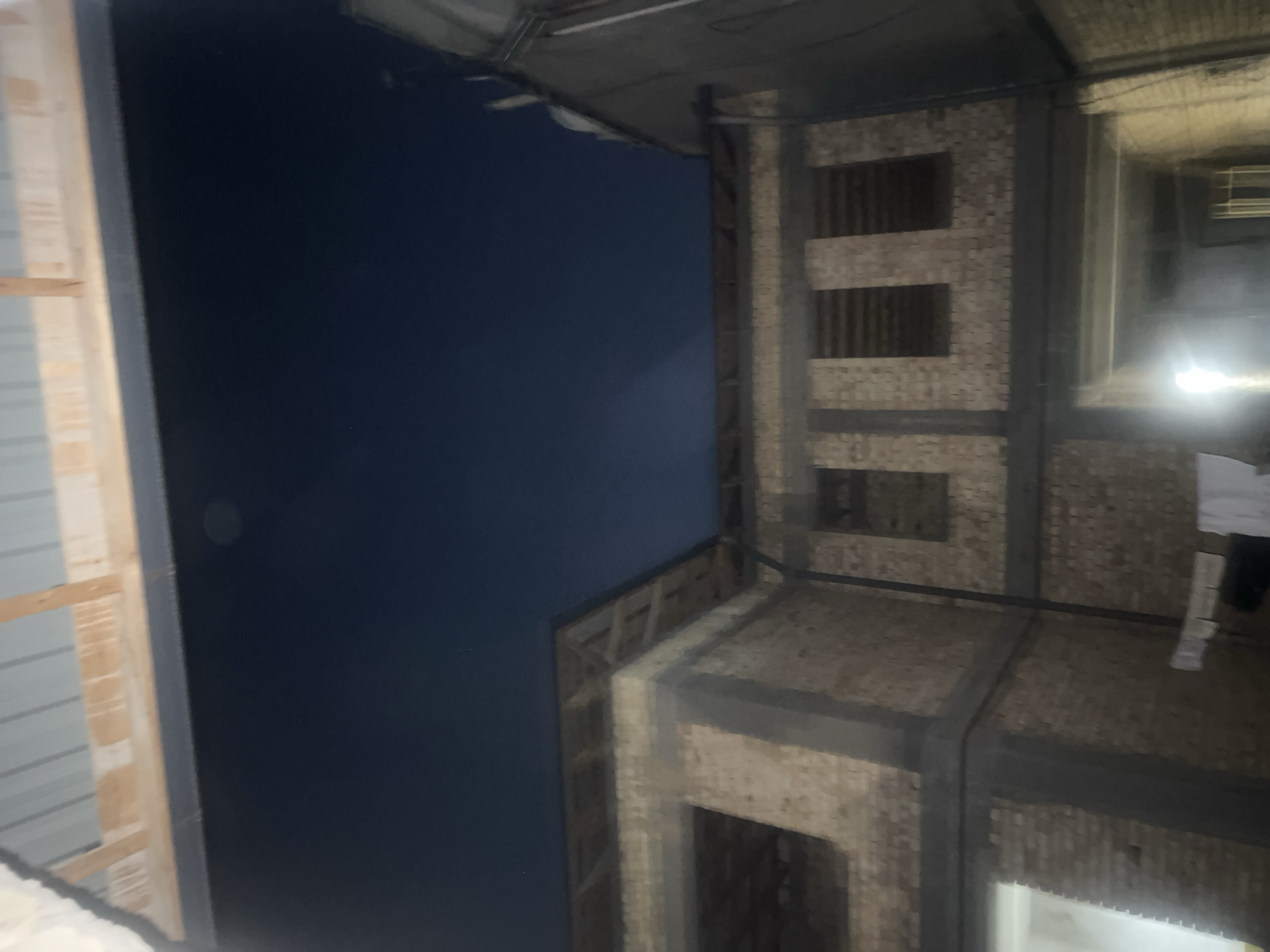 This is a photograph taken at night, capturing the exterior of a brick building under construction. The building appears to be multi-story with visible structural elements and unfinished brickwork. Openings that will likely become windows or doorways are apparent. The roof structure is exposed, showing wooden beams. Above the building, a dark night sky is visible. The overall scene suggests an ongoing construction project in an outdoor urban environment.