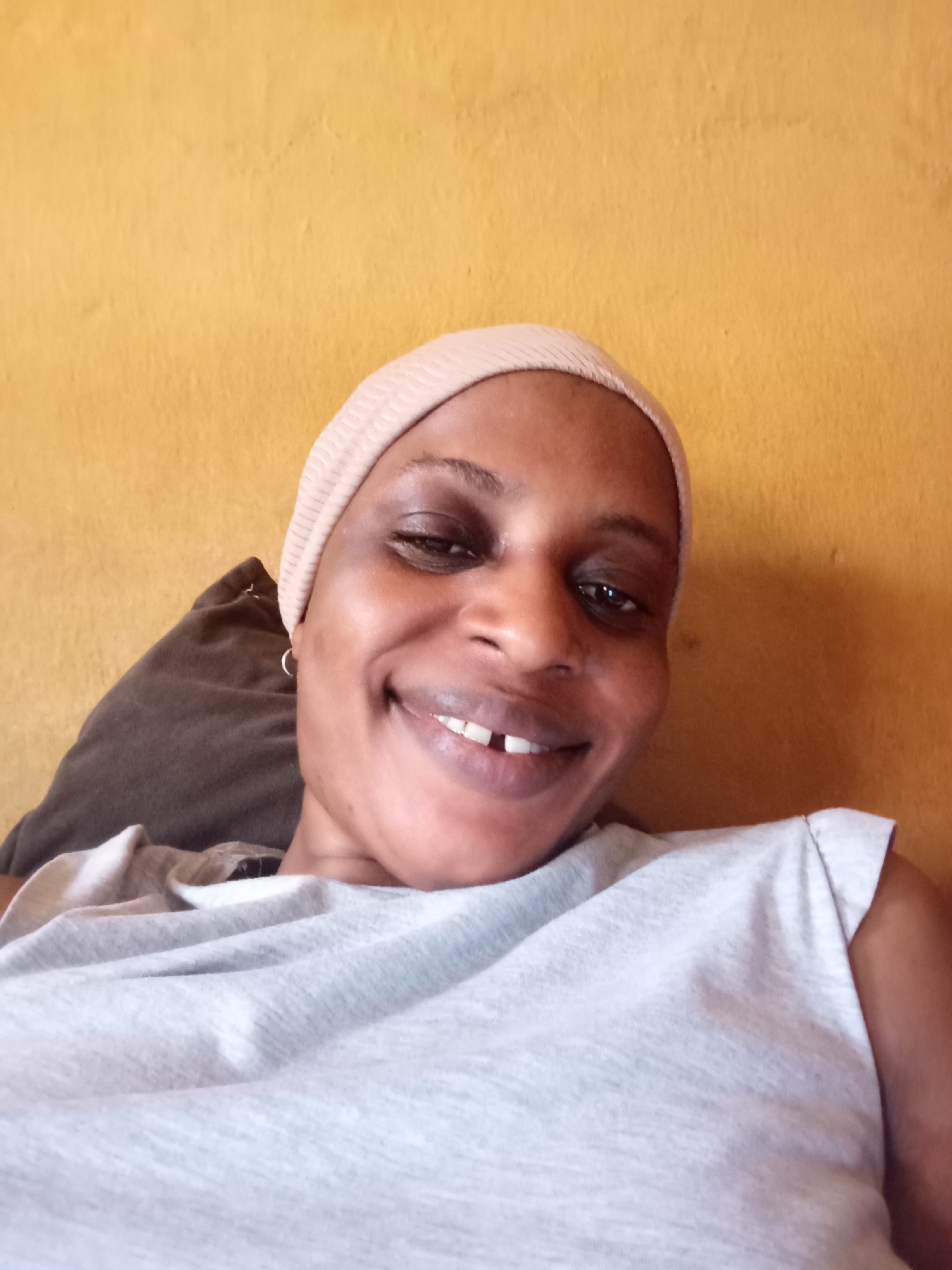 A medium-shot selfie of a dark-skinned woman smiling with a gap in her front teeth. She wears a light-pink head covering and a light-grey sleeveless t-shirt. She is leaning against a dark-brown pillow, with a yellow wall behind her. The location is reported to be Narayi, Nigeria, but there is no verifiable evidence of location in the image.