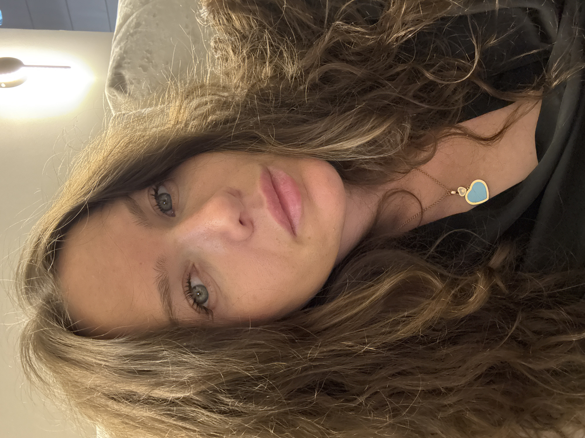 A woman with long, wavy brown hair is posing for a selfie. She has light skin, blue-green eyes, and is wearing a black top and a gold necklace with a blue heart pendant. The background is blurry and shows a light-colored wall with a wall sconce. There is no discernible information about the location in Khimki, Russia, or any other city.