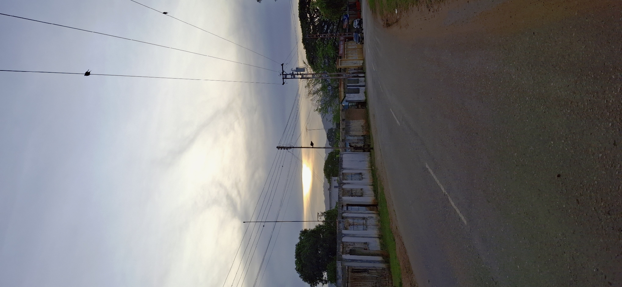 A roadside view shows a paved road curving slightly to the right under a cloudy sky with the sun peeking through in the distance. Along the left side of the road is a long, low, light-colored concrete wall with barred windows and metal gates. A large leafy tree stands behind the wall on the far left. Power lines with multiple wires crisscross the sky, and two birds are perched on them. On the right side of the road, there is a dirt shoulder, more power lines, and a building with yellow signage that reads "YOLIMAR." A couple of parked cars are visible in front of this building. The scene appears to be in Tinaquillo, Venezuela, judging by the architecture and vegetation.