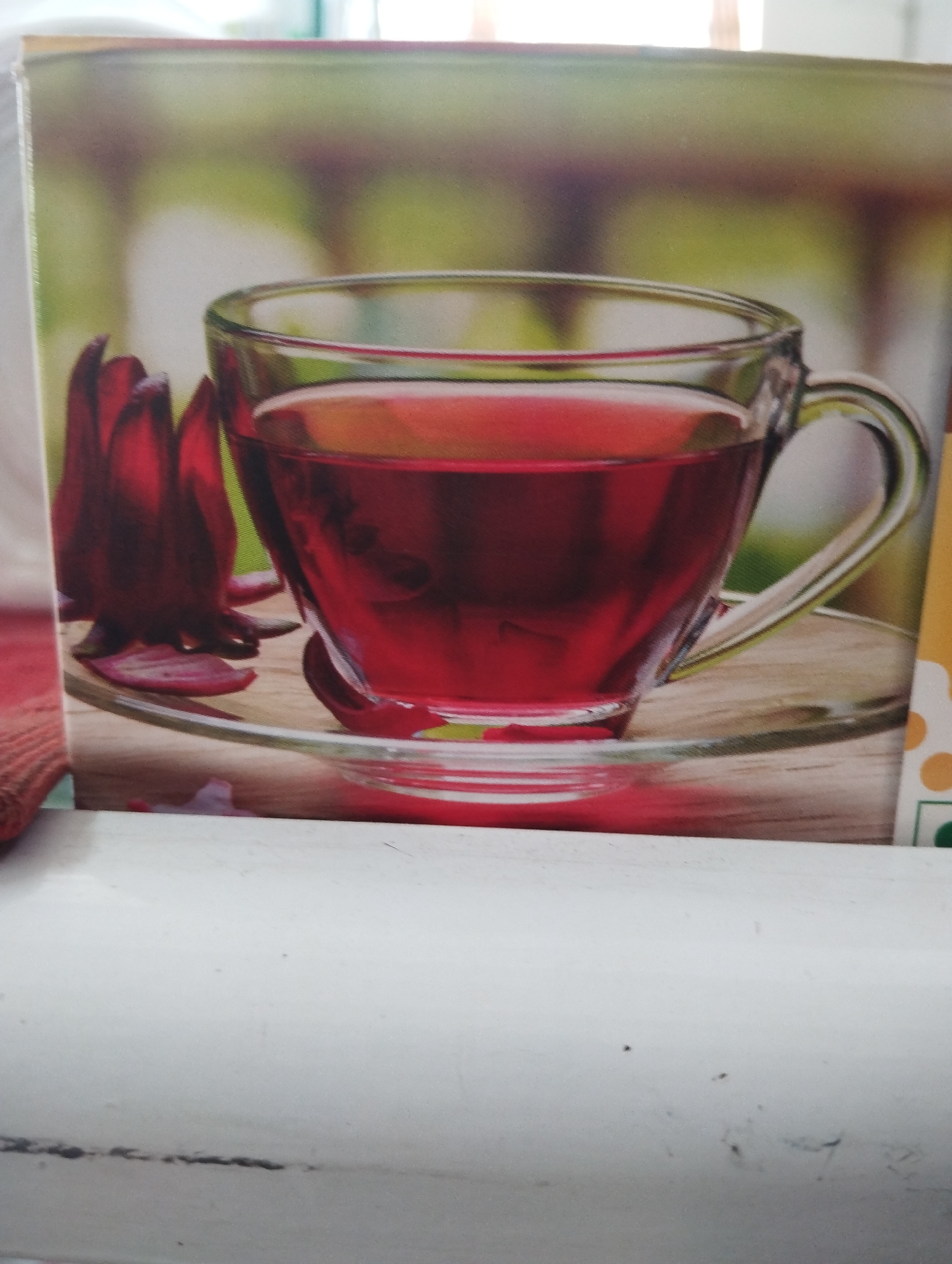 An image shows a close-up view of a glass cup of red liquid, possibly tea, sitting on a saucer. Next to the cup are a few dark red, flower-like buds. The background is blurred, with green and brown tones, suggesting an outdoor or natural setting. The image is a photograph of a printed product, likely packaging for a food or beverage. The overall scene is well-lit and the focus is on the cup and its contents. There are no people visible in the image.