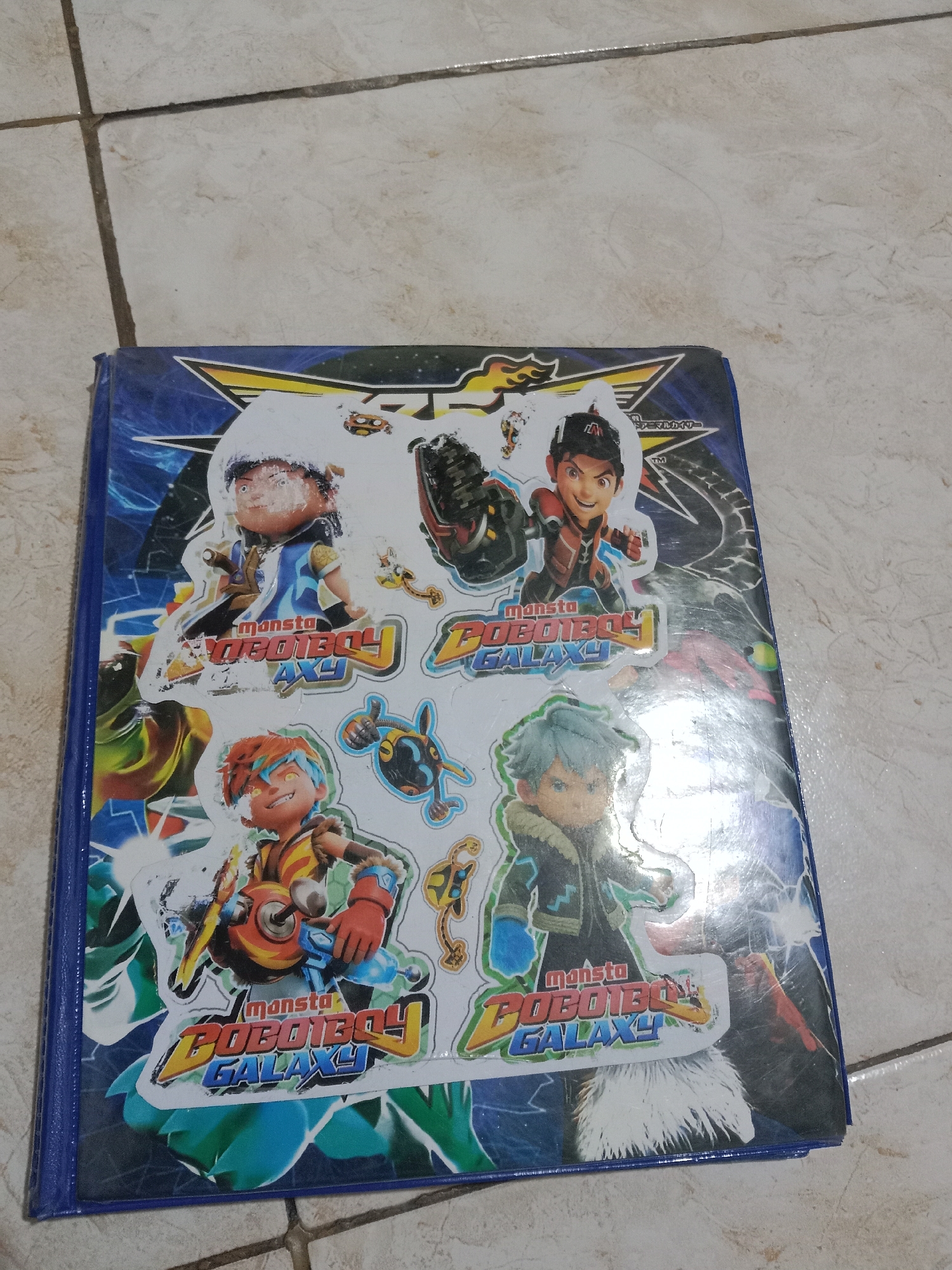 The image shows a blue binder filled with stickers featuring characters from the animated series "BoBoiBoy Galaxy." The binder is lying flat on a tiled floor. The tiles are a light beige color with darker grout lines forming a grid pattern. The binder's cover is adorned with various stickers. On the left side, there is a sticker of a character with orange hair and blue streaks, holding a weapon. To the right of this character, there is a sticker of another character with dark hair and a red outfit, also holding a weapon. In the center of the binder, there are two large stickers with the text "monsta BOBOIBOY GALAXY." Additional smaller stickers of characters and other elements are scattered across the cover. The lighting in the image is bright, suggesting an indoor environment. The location is Madiun, Indonesia.