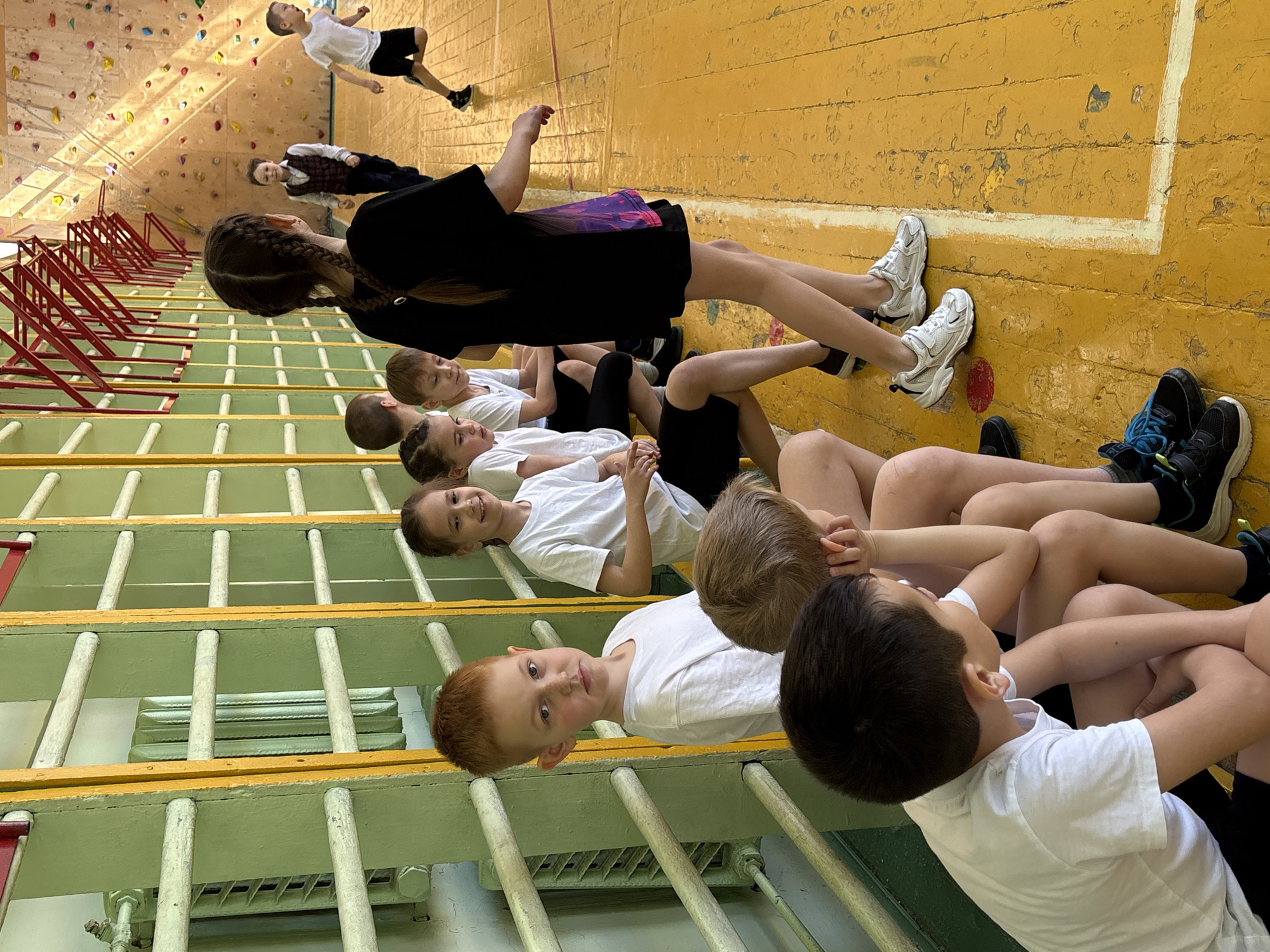 This image depicts an indoor scene in what appears to be a gymnasium or sports hall, likely in Izhevsk, Russia. The setting features a distinct yellow wooden floor with white boundary lines and a light-colored climbing wall with various holds in the background.
The primary subjects are numerous school-aged children. In the foreground and mid-ground, a group of children are seated in rows on light green wooden benches or bleachers. Most are dressed in white t-shirts and dark shorts or pants, suggesting athletic wear or a class uniform. A red-haired boy in a white t-shirt is prominently visible in the mid-ground, looking towards the camera with a calm expression. Adjacent to him, a girl with dark hair and a white t-shirt smiles towards the camera. Further down the row, another boy with dark hair is seen from behind. An older girl with long braided hair, wearing a black top and a purple/pink patterned skirt, sits closer to the camera, facing away.
In the background, two boys are visible on the yellow floor. One, in a white t-shirt and black shorts, appears to be interacting with the climbing wall, while another boy in a dark vest stands further back. Red metal frames, possibly part of gym apparatus, are present on the left side of the image. A radiator is also visible beneath the benches.
The activity taking place seems to be a physical education class, a sports club session, or a similar organized event where children are either resting or waiting for instructions. Emotions displayed by the children are generally neutral or positive, with some smiling. Faint text reading "FASHION" is visible on the side of one child's dark sneakers. The overall impression is that of a typical school sports facility.