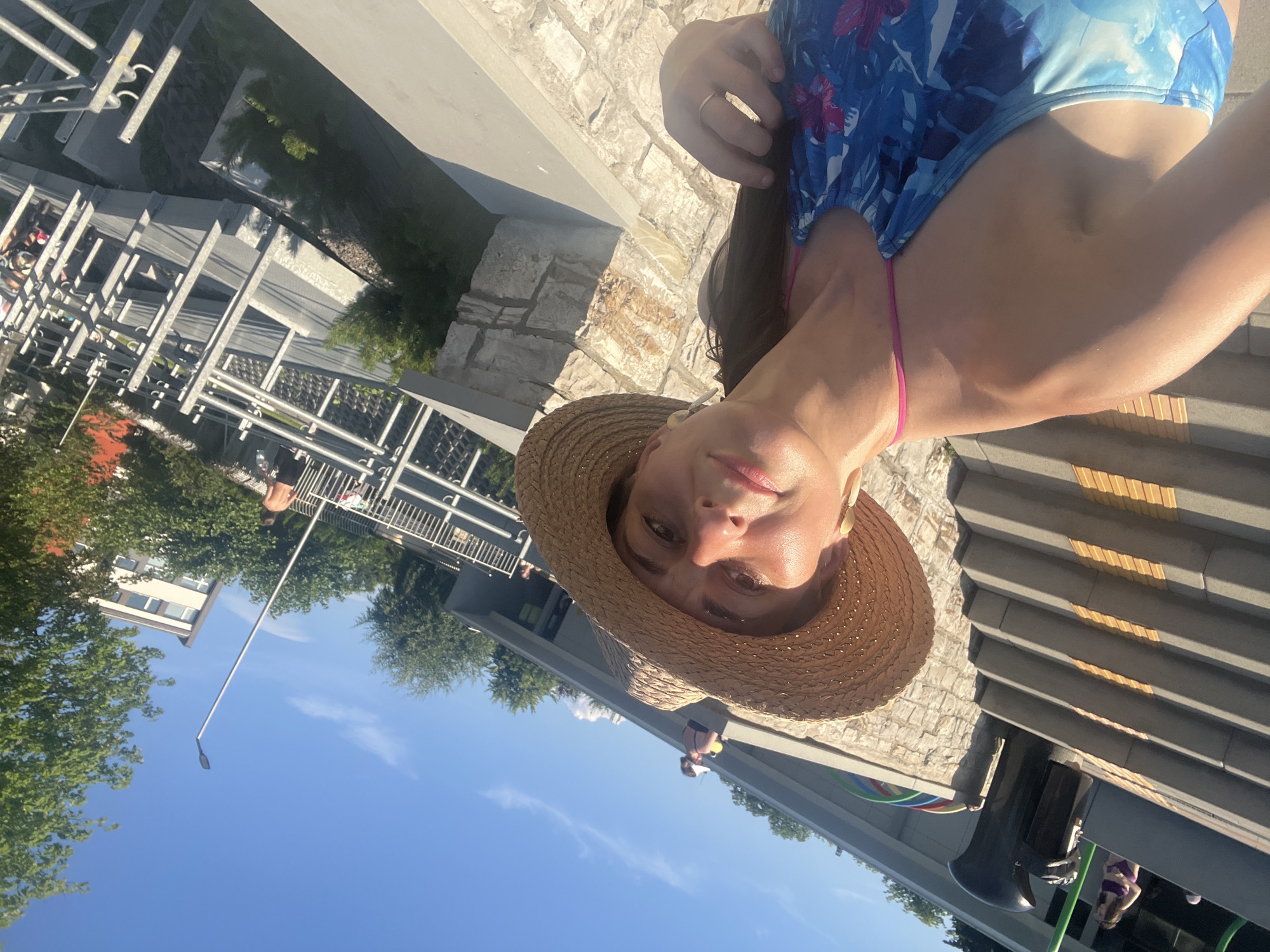 A woman wearing a straw hat and a blue floral swimsuit is taking a selfie outdoors. She has long brown hair and is wearing large earrings. Behind her, there are stone steps, a trash can, and a few other people in the background. The environment appears to be a park or outdoor recreational area with trees and buildings visible. The sky is blue and clear.