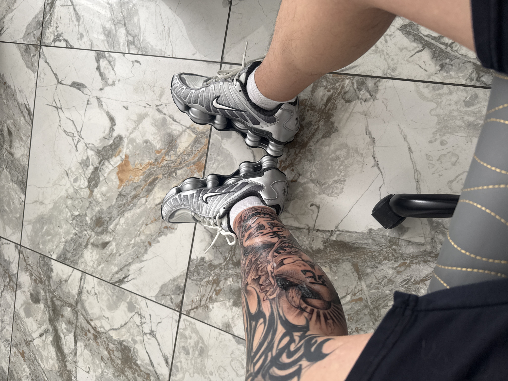 A high-angle, medium-shot photograph shows a person from the knees down. Both legs are visible, adorned with tattoos. The left leg has a prominent black and grey tattoo depicting a face and tribal designs. The person is wearing silver Nike Shox sneakers with white socks. The individual is seated, with the edge of a chair visible on the right side of the frame. The floor is covered in large square tiles with a grey and white marble-like pattern. The image lacks specific details indicating the location in Burgas, Bulgaria.