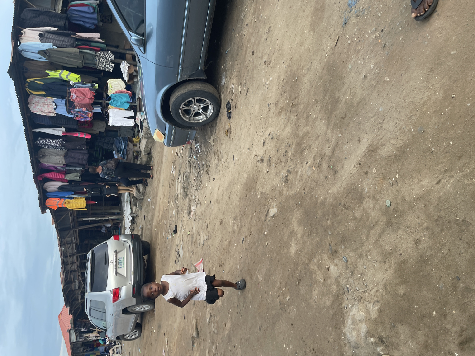 The image depicts an outdoor scene in Badagry, Nigeria. The foreground is a dirt ground with debris. A young girl in a white top and dark shorts stands on the ground. Parked vehicles, a gray sedan and a silver SUV, are visible on the left side of the frame. In the background, there's a multi-level structure with clothes hanging outside. The sky is overcast.