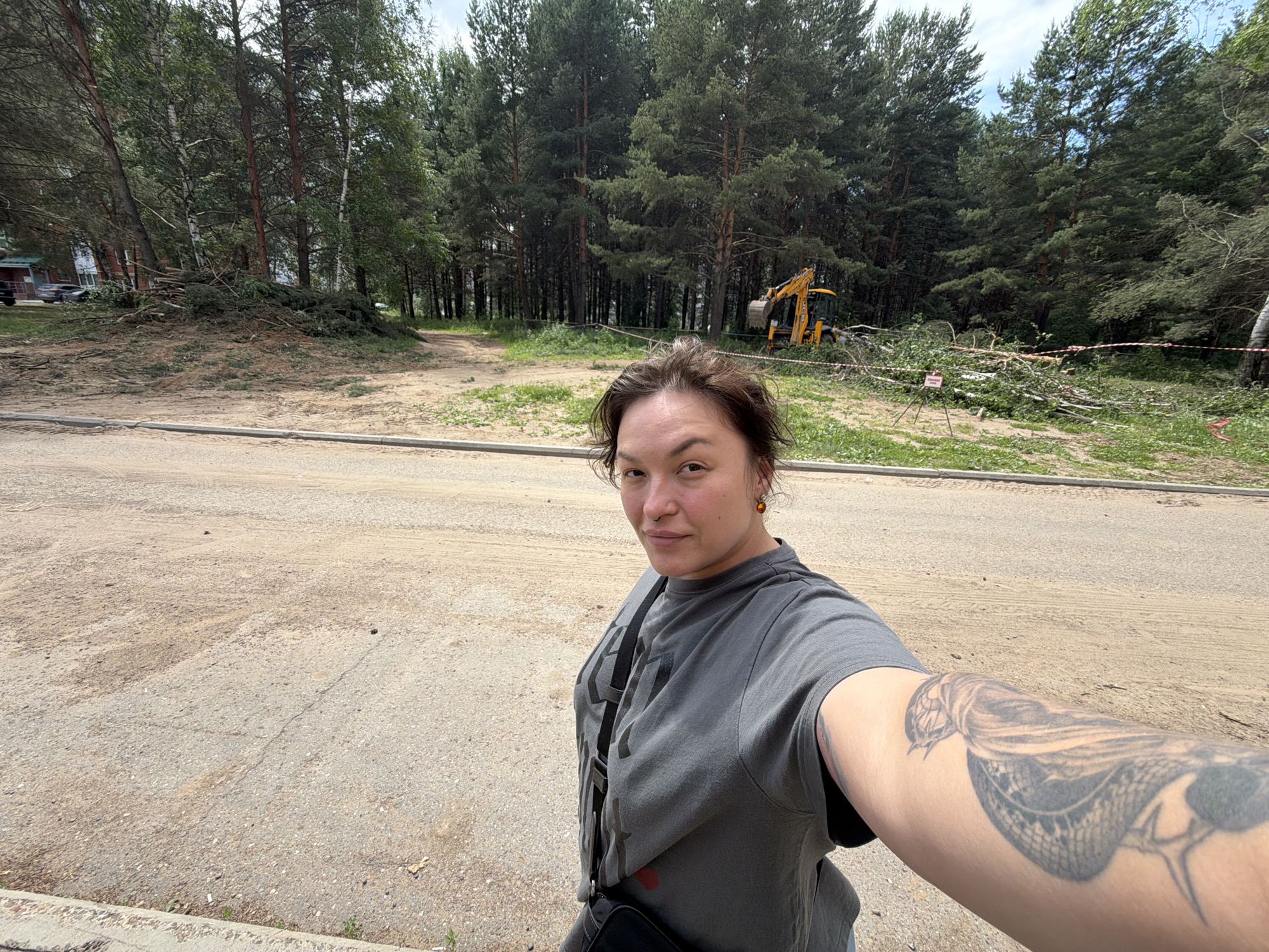 A woman is taking a selfie outdoors in Chaikovsky, Russia. She is standing on a dirt and asphalt surface, with a pile of cut trees and branches to her left and in the background. A yellow excavator is visible in the wooded area behind the cut trees. The woman is wearing a grey t-shirt and has a snake tattoo on her arm. She is also wearing a black crossbody bag. The sky is overcast and the trees are green.