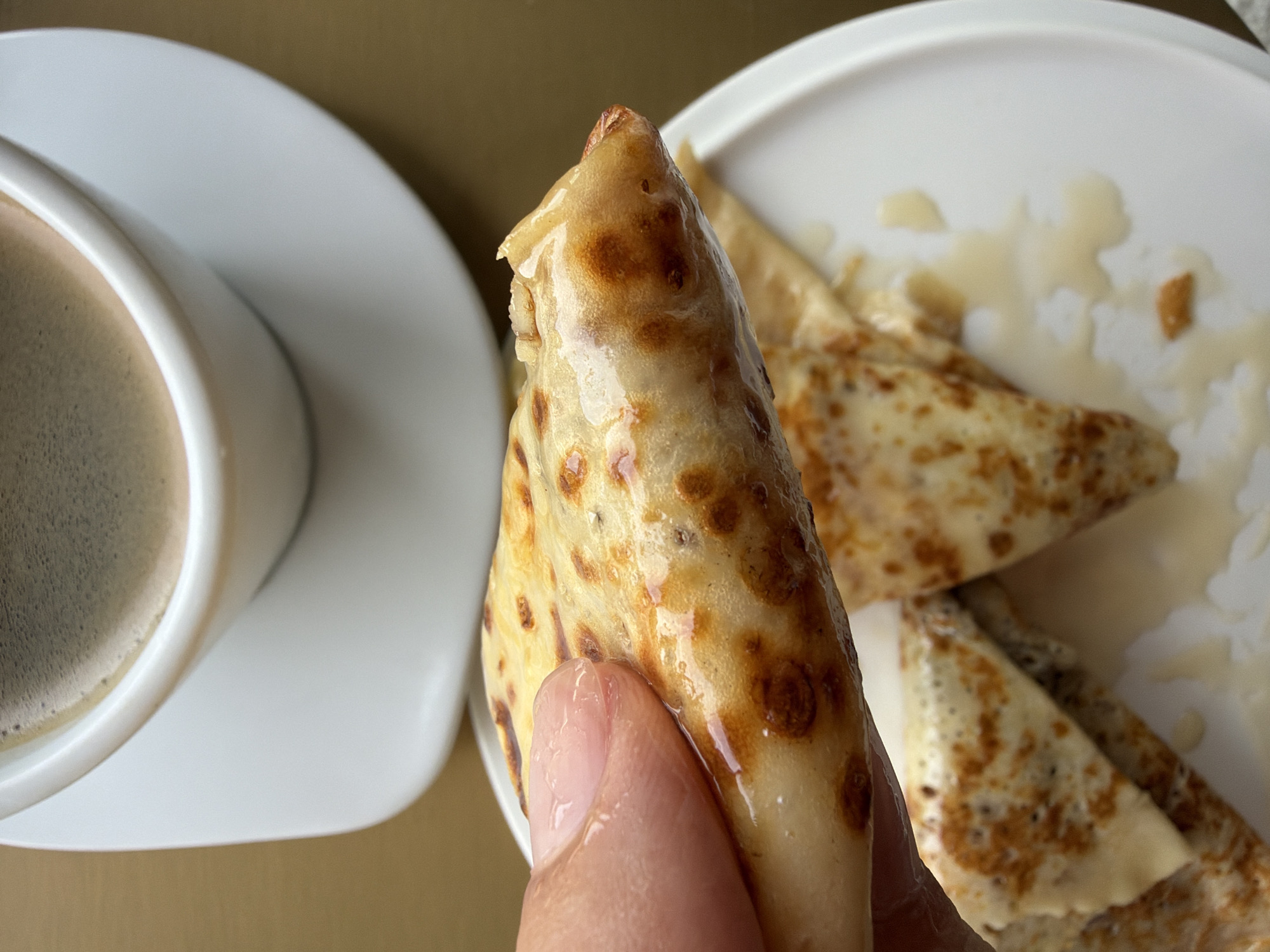 A close-up shot shows a person's finger holding a folded crepe drizzled with a light-colored syrup. The crepe is golden brown with darker, toasted spots. In the background, a white coffee cup and saucer are visible, containing dark coffee. The scene is set against a muted, earthy-toned surface. The image does not depict any specific location in Baan Bo Phut, Thailand.