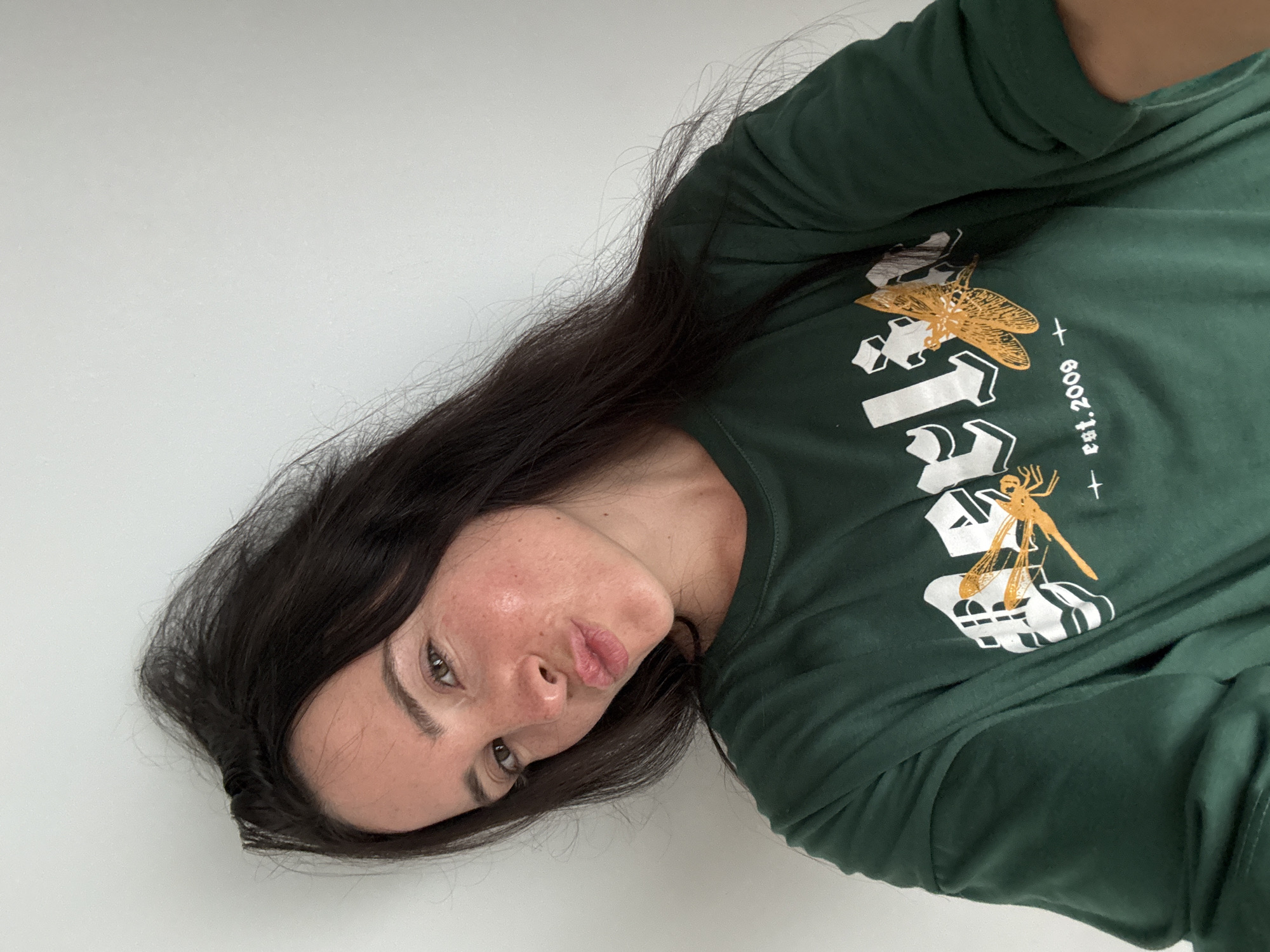 A young woman with dark hair and fair skin is looking at the camera, puckering her lips. She is wearing a dark green t-shirt with a graphic design of dragonflies and the text "est. 2009". She is positioned against a plain white wall, and her head is slightly tilted. The lighting is even, and there are no prominent shadows. The image appears to be a selfie, taken indoors.