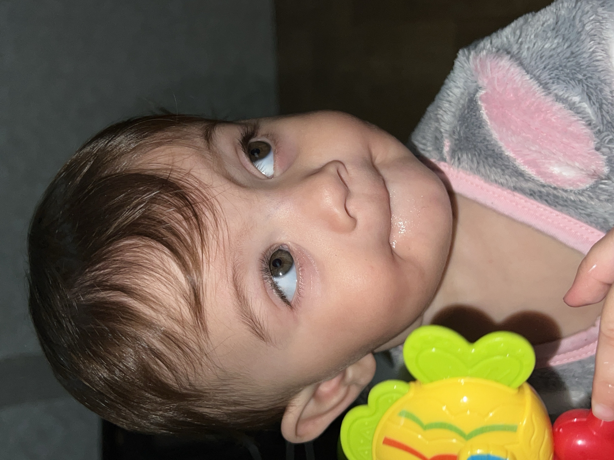 A close-up, slightly rotated image captures a baby with brown hair and eyes. The baby is wearing a grey and pink fluffy outfit that looks like a bunny suit. The baby's face is tilted, and it is looking upward. A colorful plastic toy with green, yellow, and red components is partially visible at the bottom right of the image. The background appears dark and indistinct. There is no indication in the image that the location is Elena, Bulgaria.