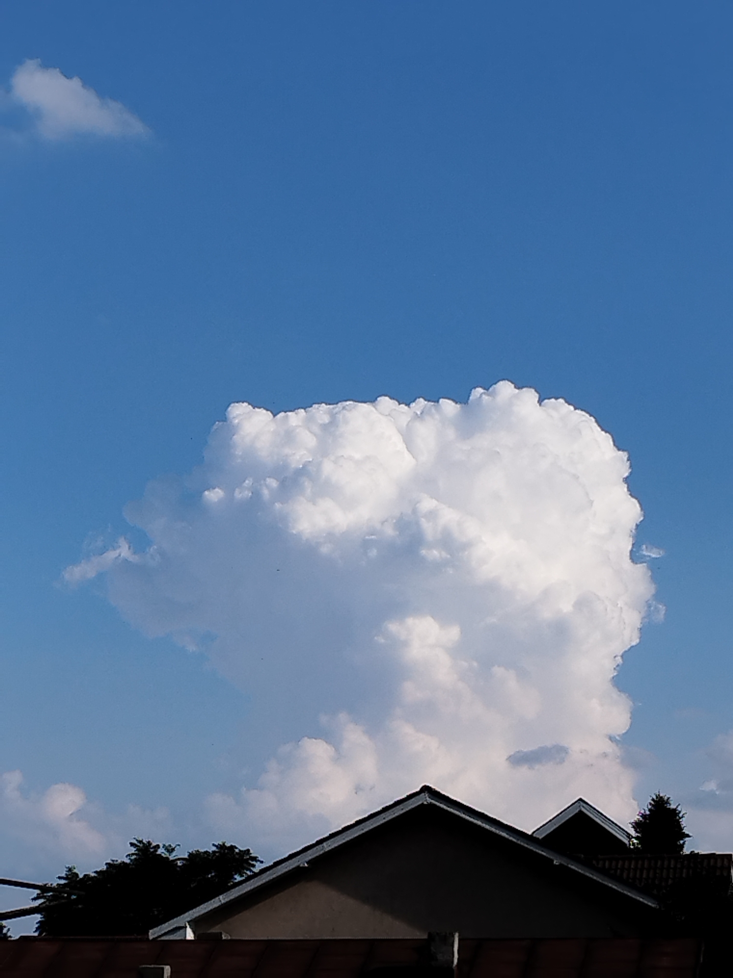 The image shows a bright blue sky with several white, fluffy clouds. A large, prominent cumulus cloud dominates the center of the frame, casting a soft shadow on the lower portion of the image. In the foreground, the dark silhouettes of rooftops and trees are visible against the bright sky. The scene appears to be an outdoor shot, possibly taken in a residential area. The location is identified as Bucharest, Romania.
