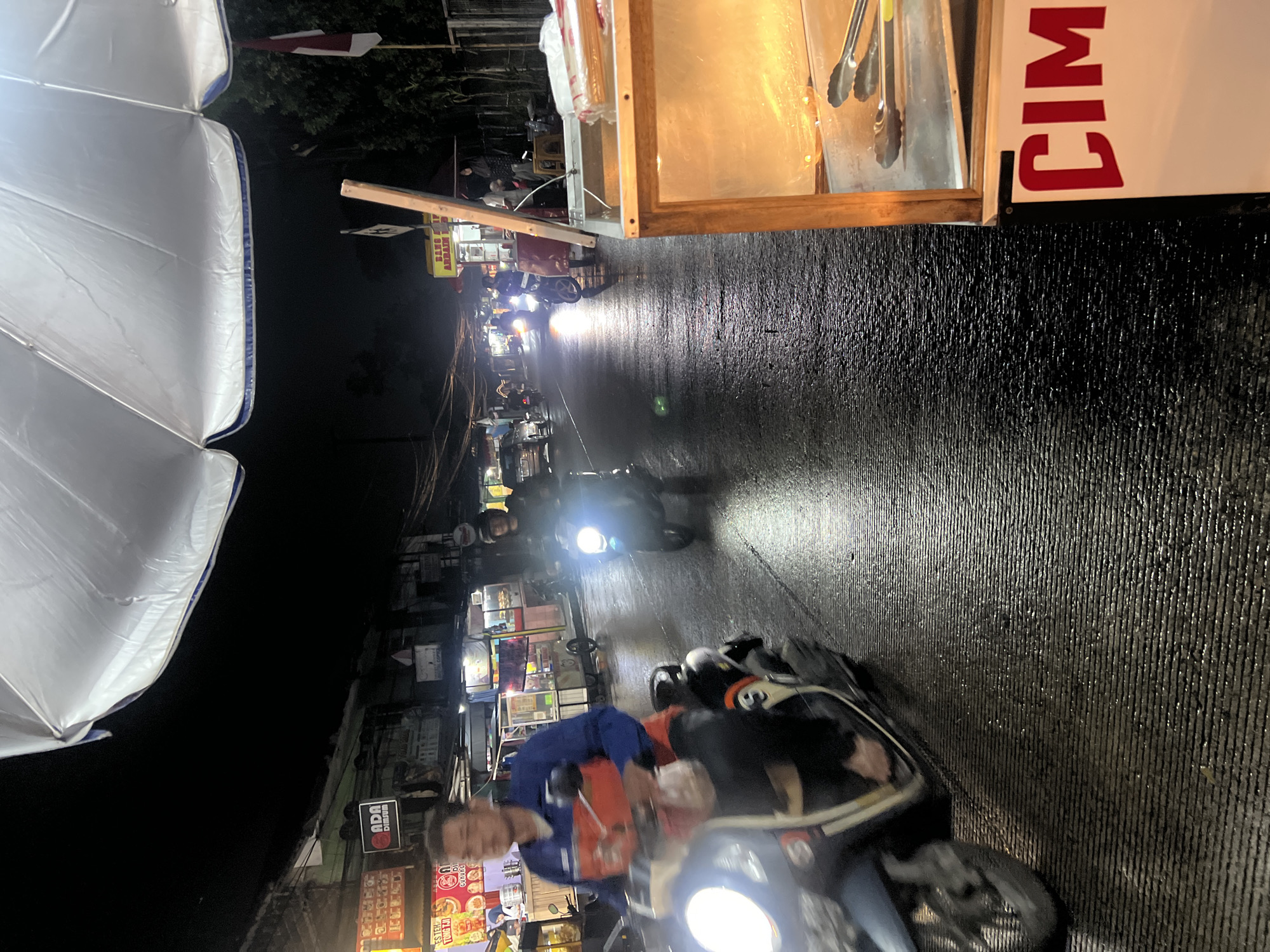 This is a nighttime street scene in Gunung Putri, Indonesia, possibly near a food stall. A person wearing a blue and red jacket is riding a scooter in the foreground, their face blurred by motion. The street is wet and reflective, suggesting recent rain. In the background, other motorbikes are visible, their headlights illuminating the road. To the right, a food stall with a wooden frame and a metal interior is partially visible, displaying what appears to be food items and tongs. A large white umbrella covers part of the upper frame. The scene conveys a sense of urban nighttime activity.
