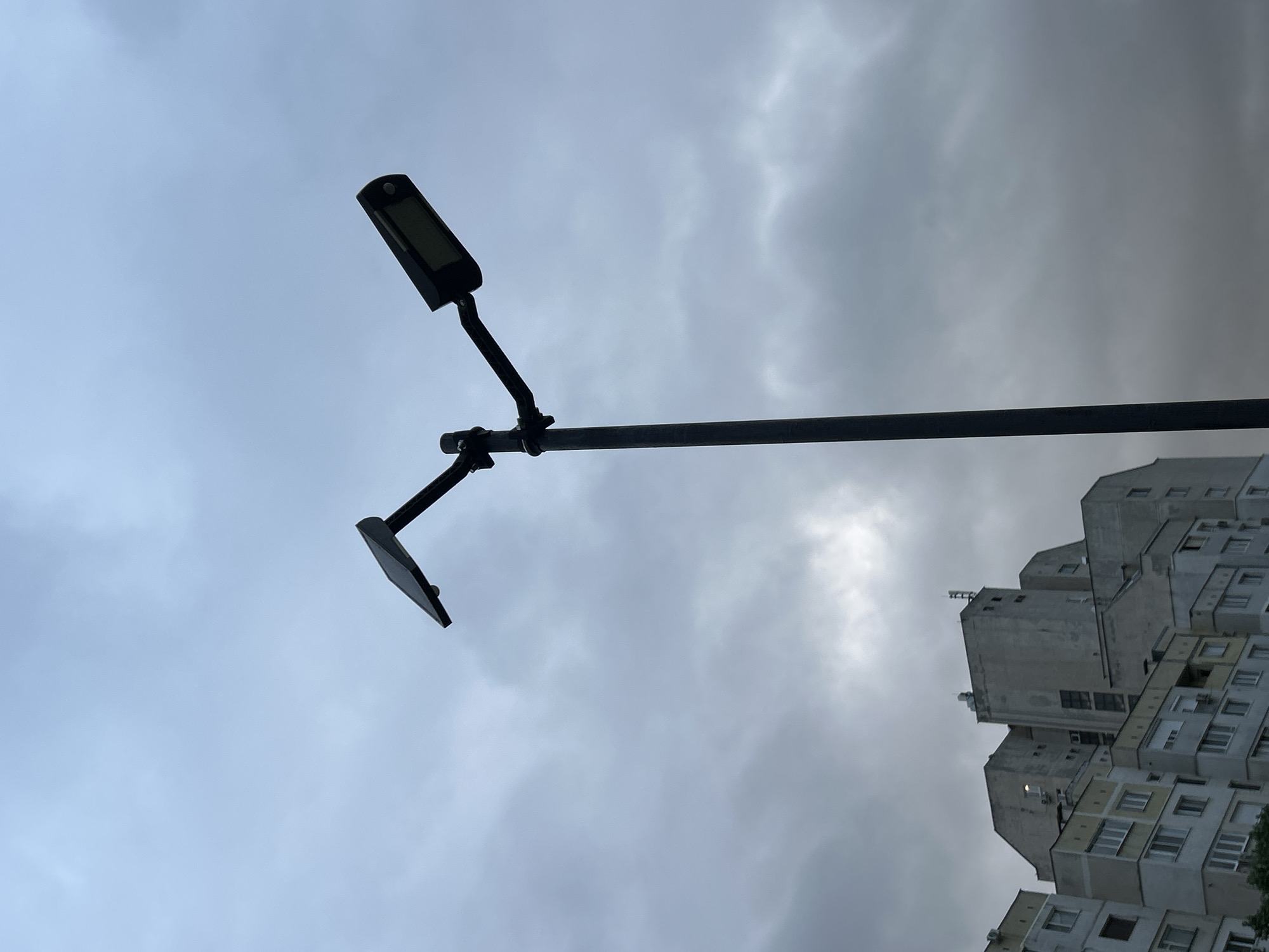 The image shows a low-angle view of a street lamp against a cloudy sky. The lamp has two lights, one rectangular and one flat. A multi-story building with a unique, stacked design is visible in the background. The location is in the city of Unknown, Unknown.