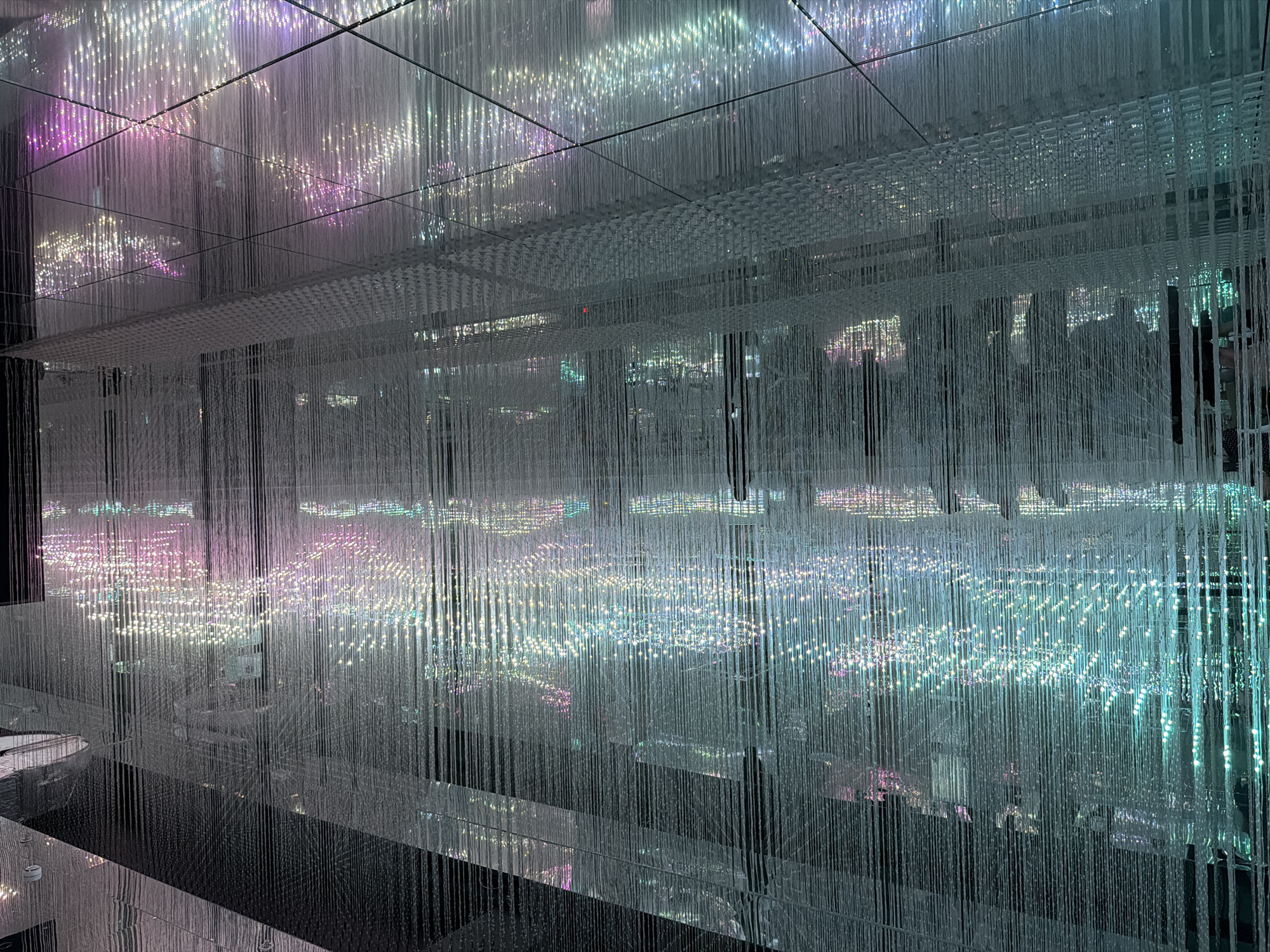 An indoor art installation features numerous hanging white threads that create a shimmering, curtain-like effect. Embedded within the threads are small lights that emit a soft glow in various colors, including teal, white, pink, and purple. The installation is illuminated by these lights and reflects off a polished, dark floor and a mirrored ceiling, creating a sense of depth and endlessness. The overall impression is one of an ethereal, dreamlike space filled with light and delicate textures. The scene is devoid of people.