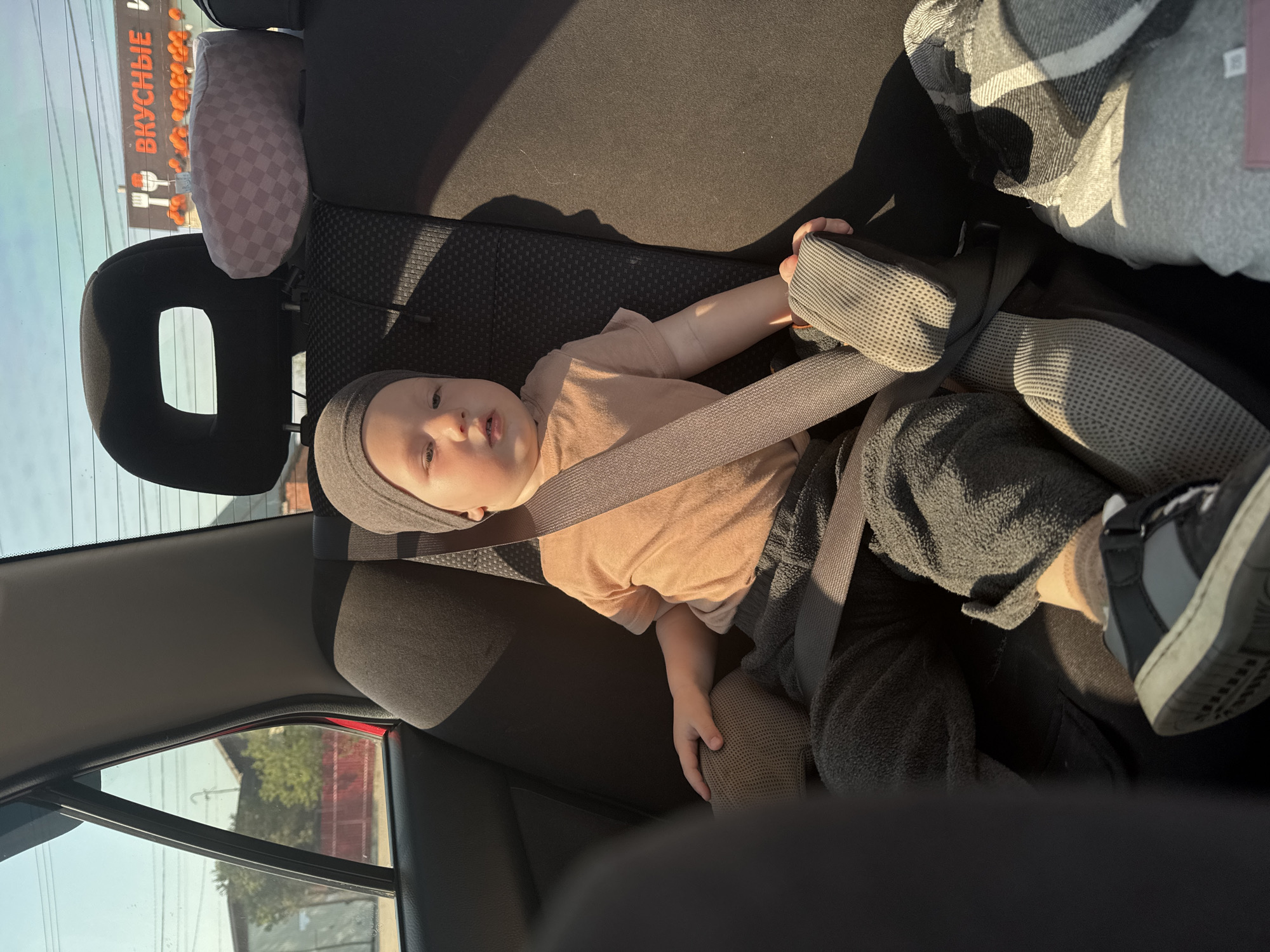 A child is lying in the back seat of a car, secured with a seatbelt. The child is wearing a light brown t-shirt, dark pants, and a gray beanie. One foot, clad in a sock and sneaker, is visible. Outside the car window, the Russian text "ВКУСНЫЕ" is visible on a sign, indicating a location in Russia. Based on this information, it is presumed to be somewhere in the city of Chaikovsky, Russia.