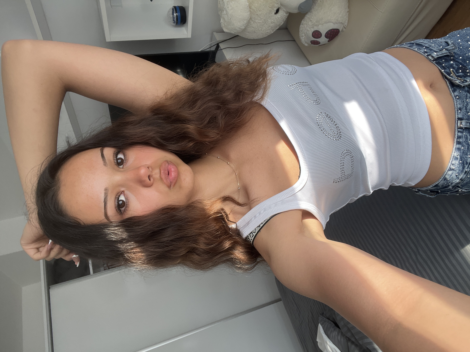 A young woman is captured in a selfie, lying down indoors with her head tilted to the left. She has long, wavy brown hair, brown eyes, and is making a playful or pouting expression with slightly puckered lips. Her arms are raised above her head, framing her face.
She is wearing a ribbed white tank top, emblazoned with the word "DREAM" in silver sparkly embellishments across the chest. A thin gold chain necklace is visible around her neck, and a black and silver patterned bracelet is on her left wrist. Her fingernails are painted white. The bottom part of her outfit consists of denim shorts or a skirt, also adorned with small silver embellishments.
The setting appears to be a brightly lit room, possibly a bedroom or living area, illuminated by strong natural light casting distinct shadows. In the background, a white shelf with a small blue and black object (possibly a mini disco ball or speaker) is visible. A light-colored, possibly beige, couch or chair is seen behind her head, along with two plush teddy bears, one large and one smaller, partially obscured. Other elements include plain white and grey surfaces, likely walls or furniture. The image is rotated ninety degrees counter-clockwise from a standard upright orientation.