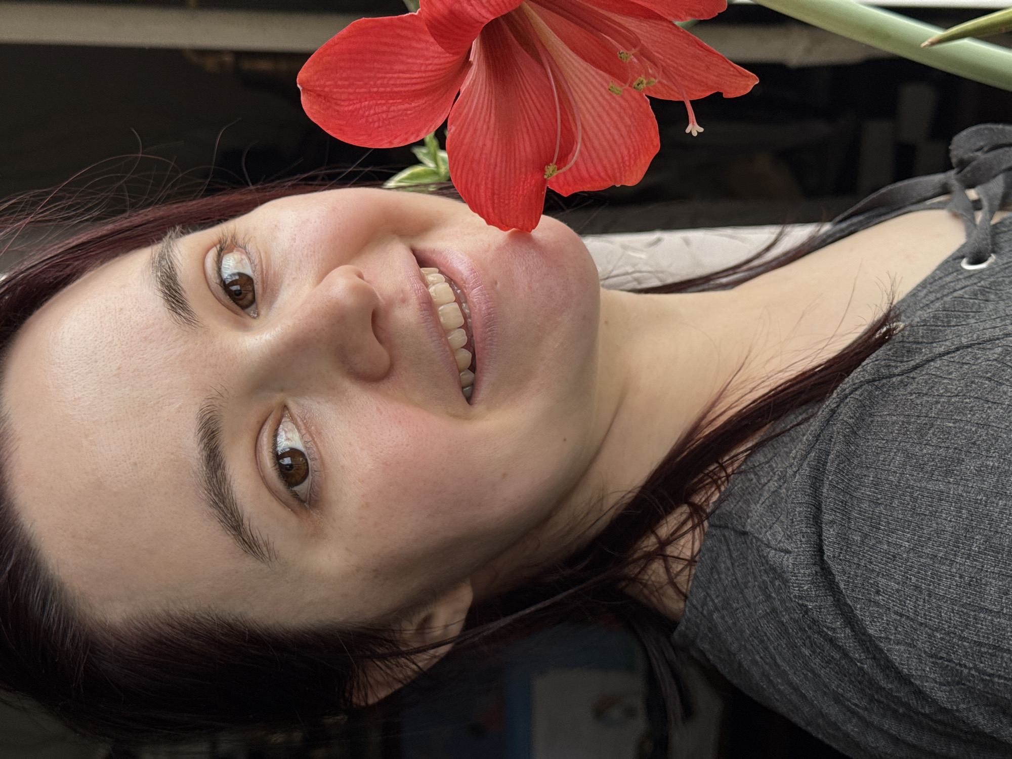 A close-up, rotated portrait features a young woman smiling broadly alongside a vibrant red amaryllis flower. The woman, appearing to be in her 20s or 30s, has fair skin, dark brown hair, and warm brown eyes that twinkle with amusement. Her wide smile reveals her upper teeth, conveying an emotion of happiness and playfulness. Her natural skin texture and well-defined eyebrows are visible.
A striking red amaryllis flower, with its distinctive petals and prominent stamens and pistil, is positioned just above her mouth and nose, seemingly touching her face. A segment of the flower's green stem is also visible. She wears a grey ribbed top with a lace-up detail on the shoulder or neckline.
The background is dark and heavily blurred, making it indistinct and suggesting an indoor setting or a shaded outdoor area. The lighting on the woman's face appears soft and natural. The image is rotated 90 degrees clockwise, giving it an unconventional and dynamic composition. No visible text is present in the image. The scene emanates a cheerful and intimate mood. The location context for this media file is Izhevsk, Russia.