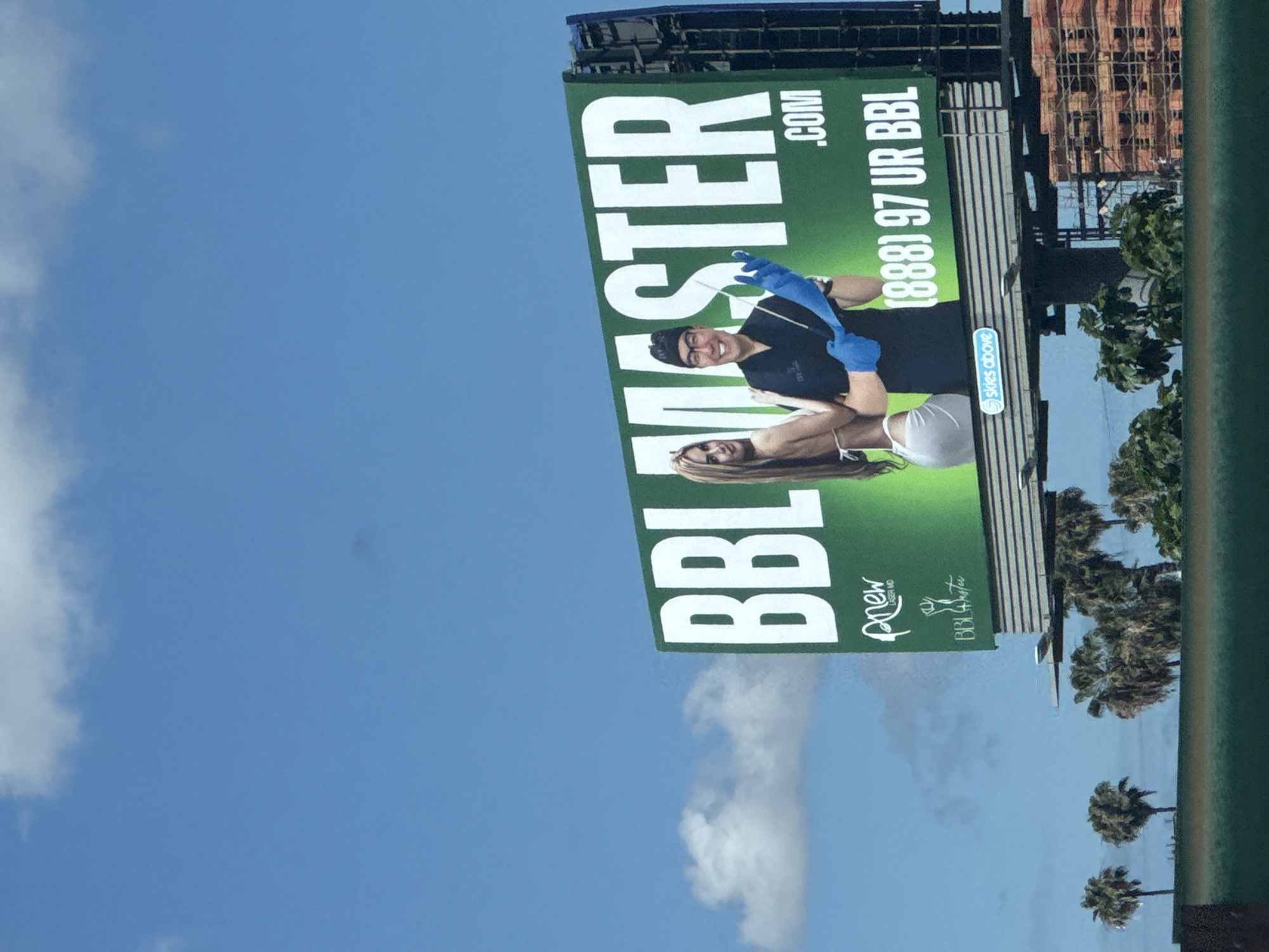 The image shows a billboard advertising a Brazilian Butt Lift (BBL) cosmetic surgery service in Los Angeles, California, USA. The billboard is vertically oriented and displays the text "BBL MASTER.COM", along with the phone number "(888) 97 UR BBL". A photo of a surgeon wearing blue gloves and smiling and a woman is featured on the billboard. The name "Renew Body & Bottox" is also displayed. Palm trees and a building under construction are visible in the background, along with a clear blue sky.