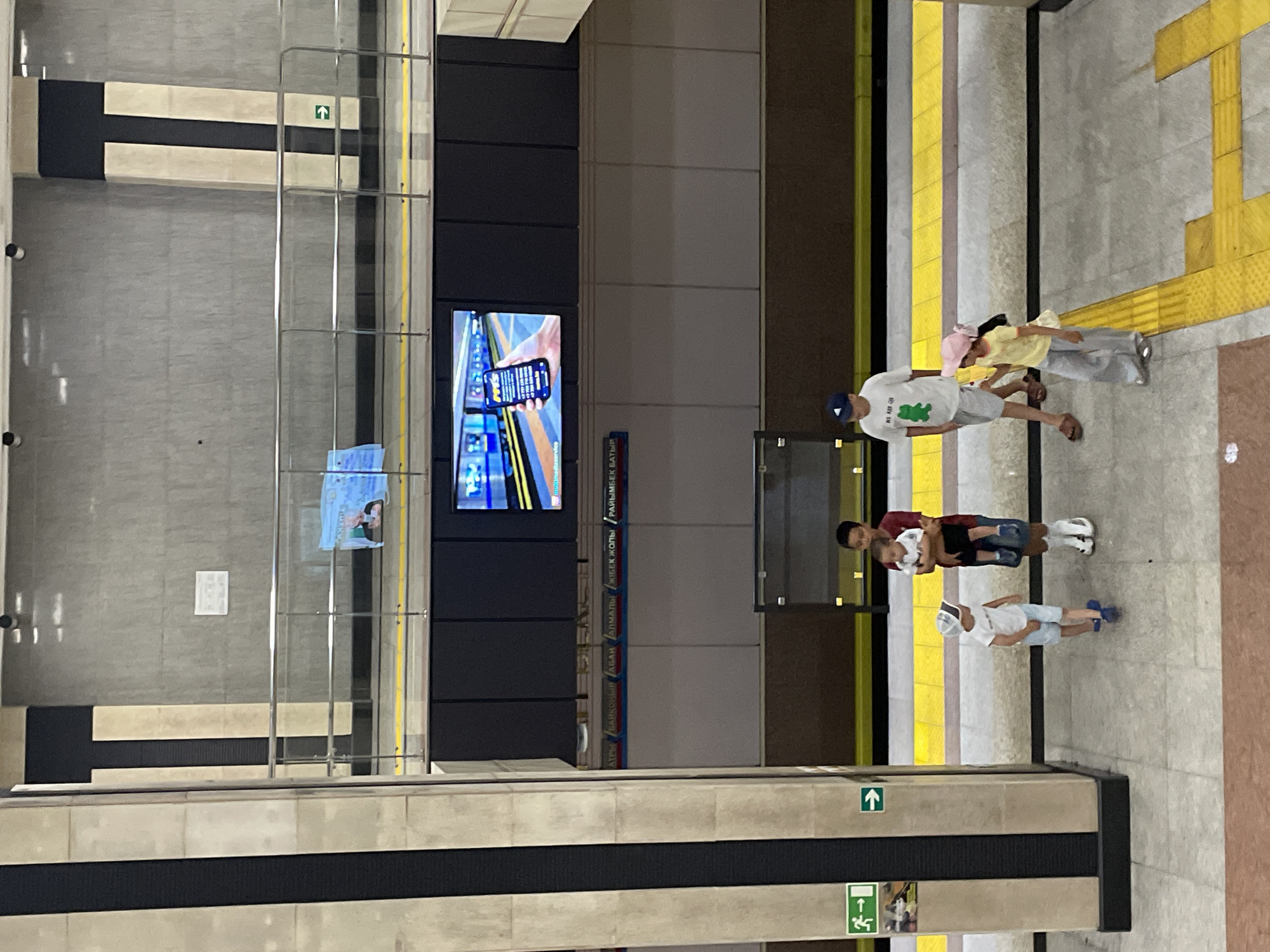 The image depicts a scene at the Almaty Metro station, likely during daylight hours. The perspective is from a higher vantage point, looking down onto a platform. In the foreground, on the tiled platform floor, are four people. A man, wearing a white t-shirt with a green frog graphic, light shorts, and a blue baseball cap, walks from left to right. Beside him, a young girl in a yellow t-shirt and a pink hat, wearing gray pants and sandals, walks towards the right. To the left, a younger boy in a white t-shirt and light blue shorts, also wearing a cap, walks towards the right. Further to the right, a man is holding a toddler. The man is wearing a dark red shirt and dark shorts, and the toddler is in a white outfit. In the background, above the platform, a large screen displays an advertisement showing a hand holding a smartphone with a subway map on it. Below the screen, station names in Cyrillic script are visible on the wall. The station features yellow safety lines along the platform edge and a clean, modern aesthetic with concrete and metal railings. Green directional arrows are visible on the walls, indicating exits.
