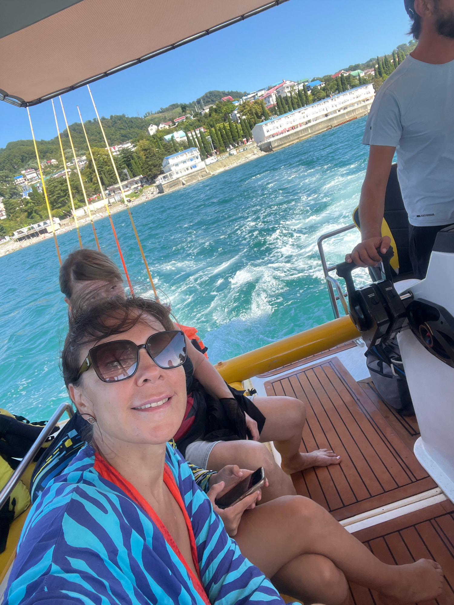 A medium shot shows a woman with sunglasses and a blue and turquoise patterned robe taking a selfie on a boat in Sochi, Russia. She is smiling and holding a smartphone in her right hand. Behind her, another person, likely a man, is operating the boat's controls, with his hand on the throttle. A second person is visible sitting behind the woman, with their hair obscuring their face. The boat has a yellow railing and a wooden deck. In the background, the coastline of Sochi is visible, with buildings, trees, and the sea. The water is choppy and a wake is visible behind the boat. The sky is clear and blue. The overall impression is of a recreational boat trip on a sunny day.