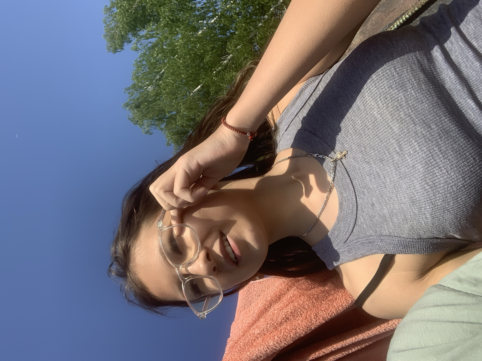 The photograph shows a young woman wearing glasses, a grey ribbed tank top and a red bracelet, outdoors in what appears to be a sunny environment. The photo is taken at an angle, looking up at the woman. A blue sky and green foliage are visible in the background. The location is not indicated by the image.
