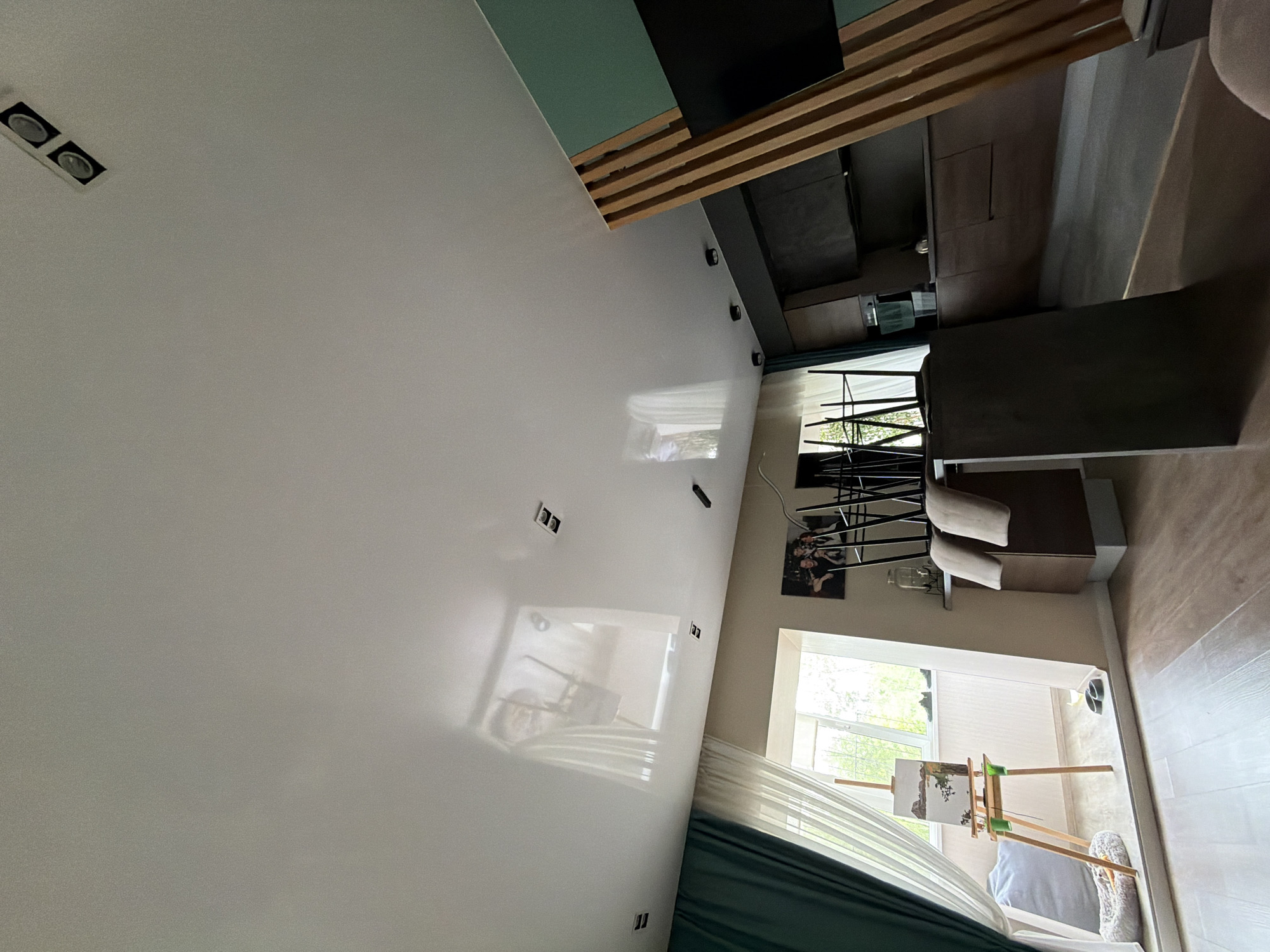 The image depicts an interior room. The room is painted white. A window lets light into the room. An artist's easel is positioned near the window. A painting on the easel shows a landscape. The room also features a couch, chairs, and a lighting fixture. The location is not specified.