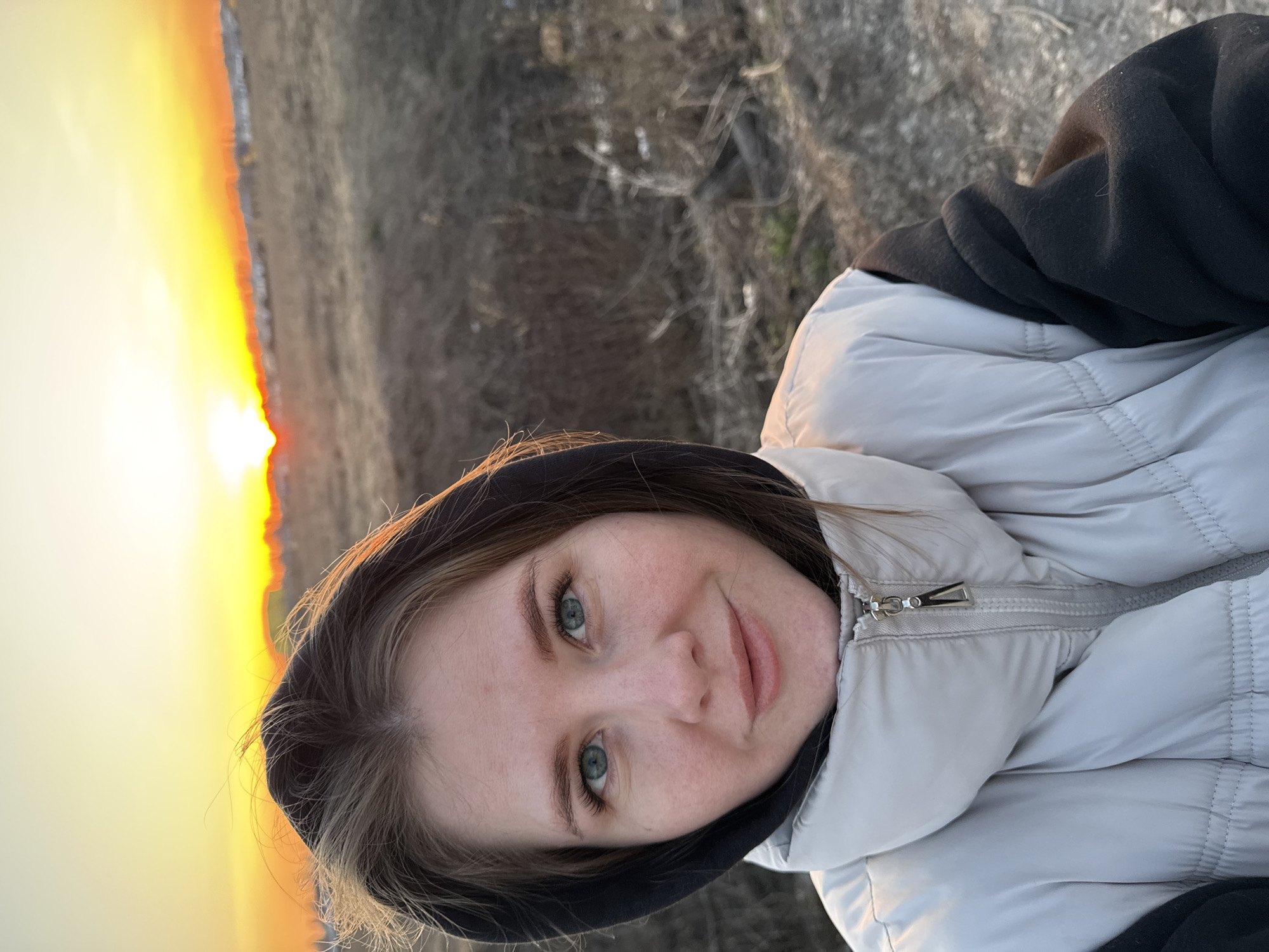 A woman is pictured outdoors during sunset in Горный, Russia. The horizon line is visible behind her, with a vibrant orange sunset. She is wearing a black hooded sweatshirt underneath a light-colored puffer jacket, the zipper of which is visible. Her face is angled towards the camera. The background appears to be a field.