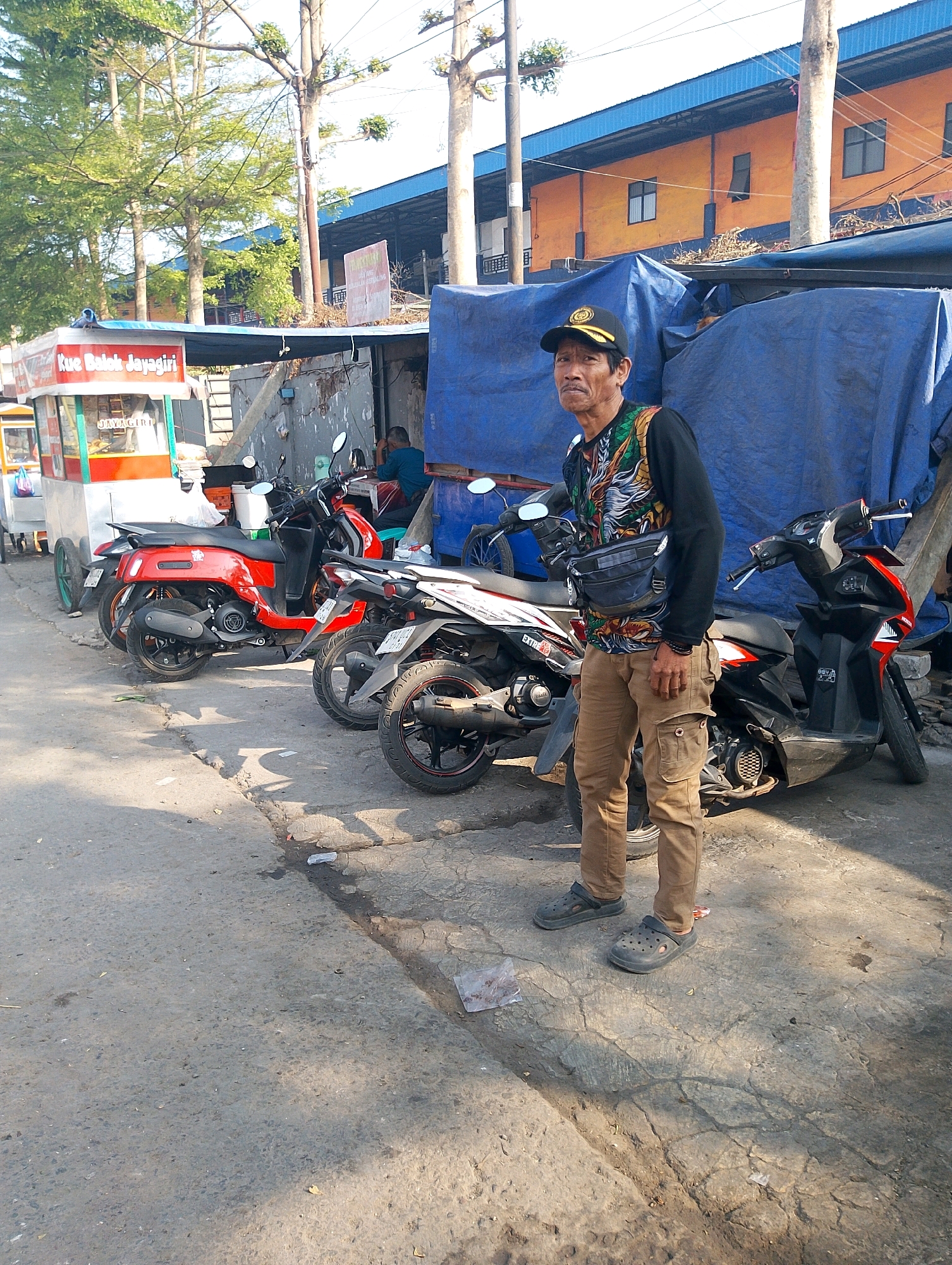 An Indonesian man wearing a black baseball cap, a long-sleeved shirt with a dragon design, khaki cargo pants, and black clogs stands on a cracked asphalt road in Lembang, Indonesia. He is positioned in the foreground, facing the camera, with a dark waist bag slung across his chest. Behind him, several motorcycles are parked along the road, some red and some black and white. A food stall with the sign "Kue Balok Jayagiri" is visible to the left. The background shows buildings with orange and blue facades, and trees with green foliage. The scene appears to be a street market or roadside commercial area.