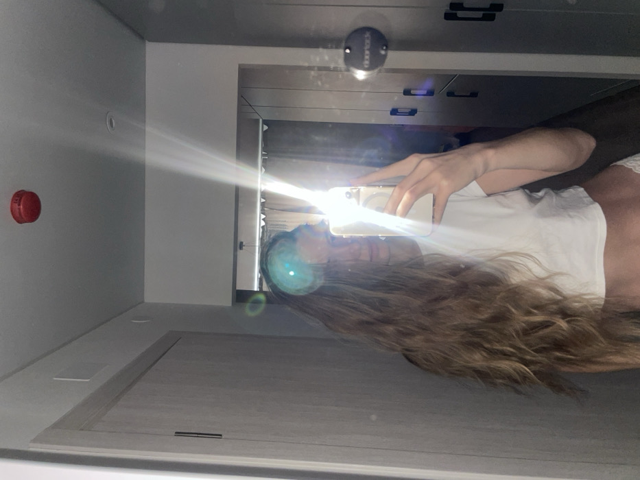 The image displays an indoor scene, located in the city of Khimki, Russia. It is a mirror selfie depicting a person with long, wavy, light brown hair. The person wears a white short-sleeved crop top, and white lace-trimmed undergarments are partially visible. A light-colored smartphone is held by the person's right hand, extended to capture the reflection. A significant lens flare, originating from the phone's light source, obscures part of the person's face and the central area of the image.
The reflected environment includes a white ceiling with a red circular smoke detector and a recessed light fixture. To the left, a light-colored wooden door with a dark vertical handle is visible. To the right, a series of dark gray cabinets or wardrobe doors with dark horizontal handles are present. A circular, dark object labeled "Doornack" is affixed to one of these doors. In the background, beyond an open doorway, a room with a dark curtain and a minimalist light fixture is partially discernible.