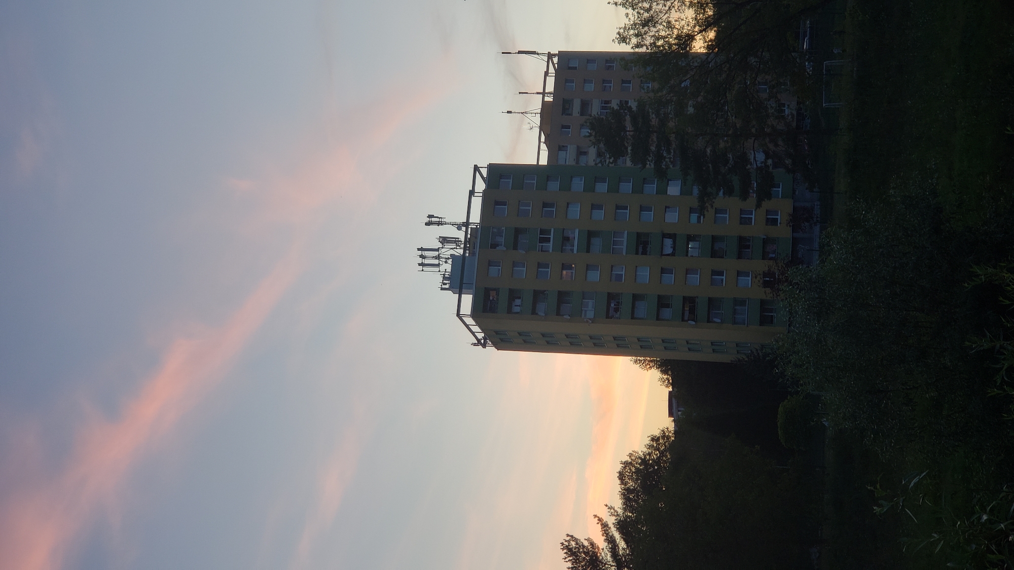 The image captures a twilight scene in Prešov, Slovakia, featuring two tall residential buildings with cellular antennas on their roofs. The sky is a gradient of soft blues and pinks, with wispy clouds streaked across it. The buildings are primarily yellow and green, with numerous windows visible. In the foreground, dark foliage and trees obscure the lower portion of the scene, hinting at a park or wooded area adjacent to the buildings. A sports field with goalposts is partially visible on the right side of the image, suggesting recreational space within the urban environment. The overall atmosphere is serene and suggests the end of a day in a residential part of the city.