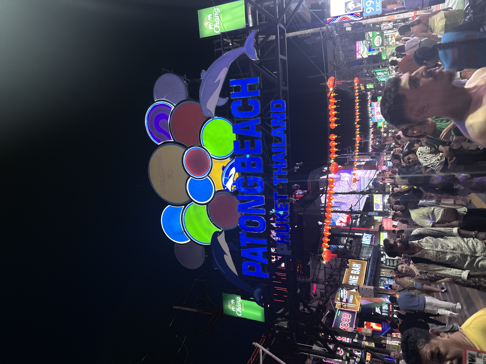 A nighttime, low-angle photograph captures a crowded street scene in Patong Beach, Phuket, Thailand. An illuminated archway dominates the upper third of the image, featuring the words "PATONG BEACH" in large blue capital letters. Above this, "WELCOME TO" is displayed on a brown circular element, flanked by numerous colorful circular lights in blue, green, purple, and red. Two stylized blue dolphins are integrated into the archway design, appearing to swim towards the center. Below "PATONG BEACH," the text "PHUKET THAILAND" is also visible in blue. On either side of the archway, large green rectangular banners with the "Chang Mineral" logo are suspended.
Below the archway, the street is densely packed with pedestrians and lined with brightly lit commercial establishments. Numerous neon signs are visible, including "HOLLYWOOD," "ONE BAR," "PHARMACY," and "MYST." A string of red Chinese lanterns stretches across the street overhead. A large digital display on the right shows "99 EXC." A complex network of overhead wires is present throughout the scene.
The foreground is occupied by a diverse crowd of people walking in various directions. Prominently featured individuals include a man in a yellow shirt partially visible on the far left, a man with a beard and mustache wearing a patterned long-sleeved shirt and a backpack looking forward, and a woman in a patterned dress beside him. On the far right, a man with a fade haircut, shirtless or in a light-colored top, is seen in profile looking right. The overall environment is bustling and heavily illuminated by artificial light sources.