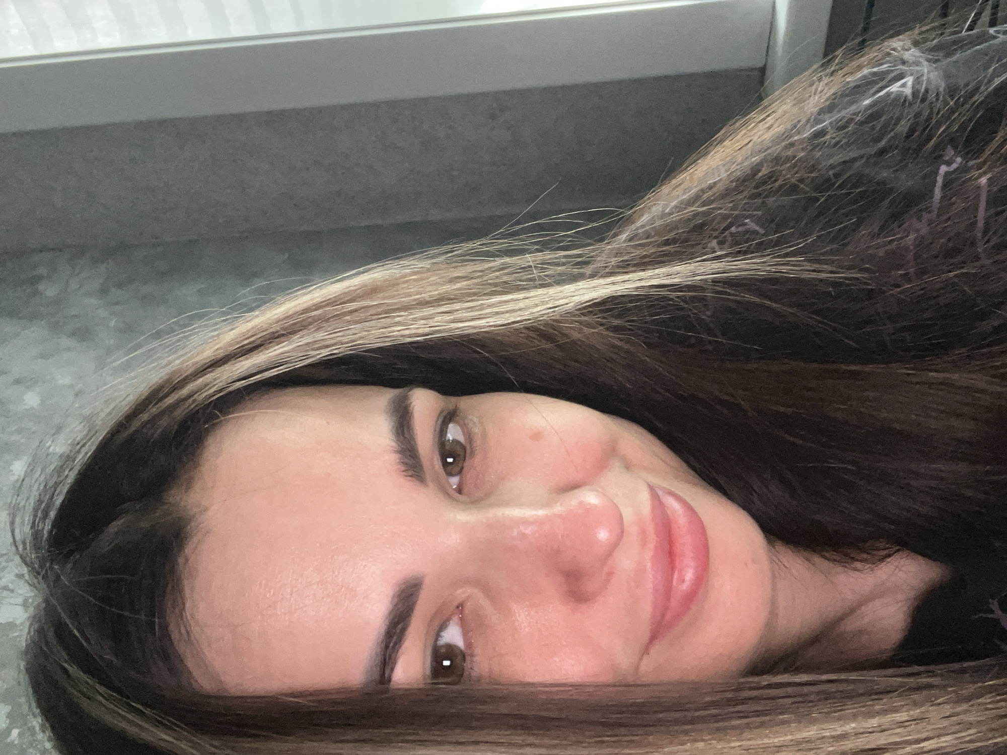 A close-up, canted portrait of a woman with long, brown hair with blonde highlights. Her eyes are brown, and she has a small mole on her cheek. She is smiling slightly. The background is a grey, textured surface with a white horizontal line at the top of the frame. Location: Unknown, Unknown.