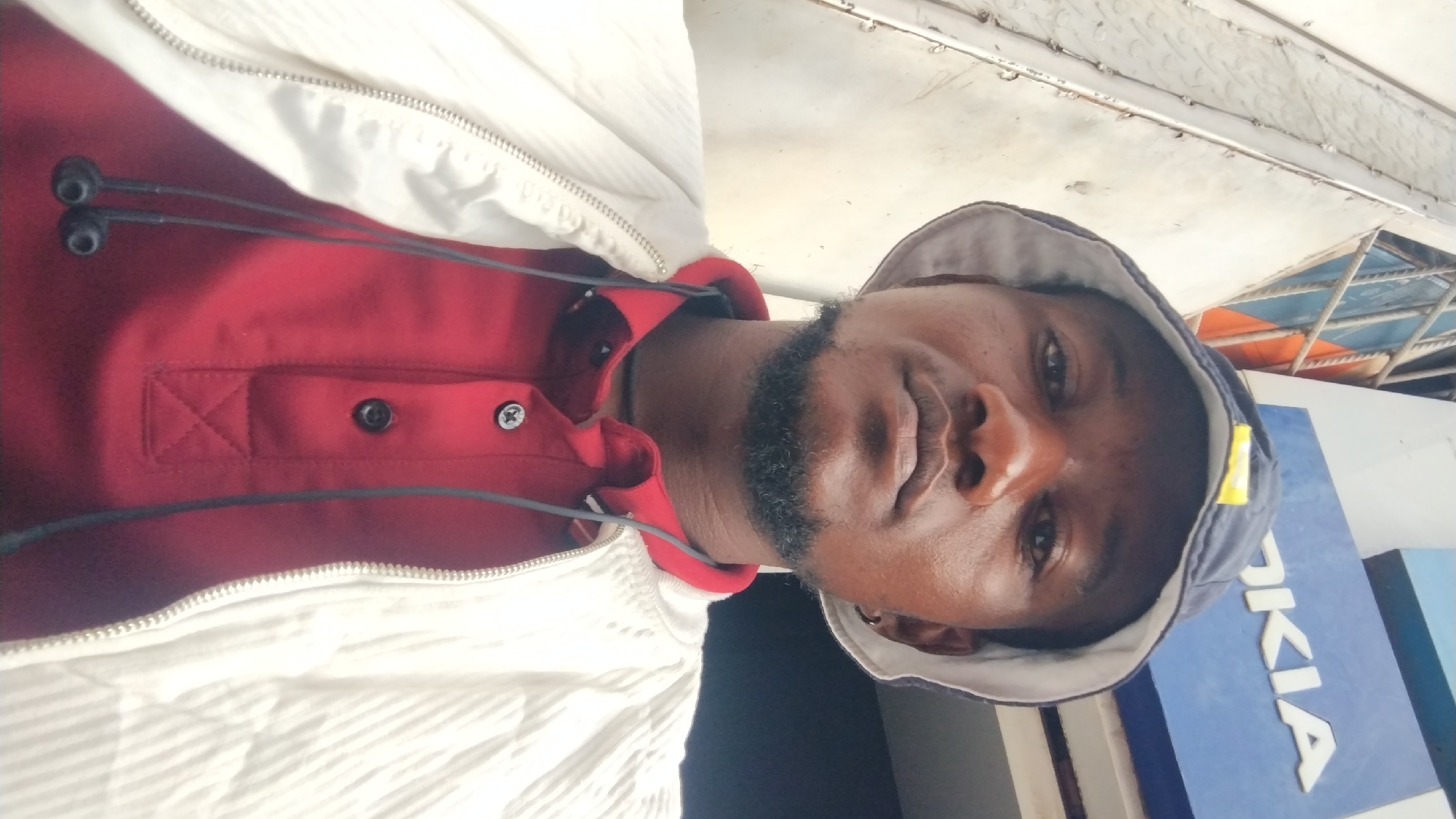 A man wearing a red shirt, white jacket, and a bucket hat is posing for a picture. He is standing in front of what appears to be a Nokia advertisement, indicating the possible location is Jos, Nigeria, given the presence of the brand in that region. The man has dark skin and a beard. He has headphones hanging around his neck. The background is a mix of architectural elements. The picture is taken from a slightly low angle.