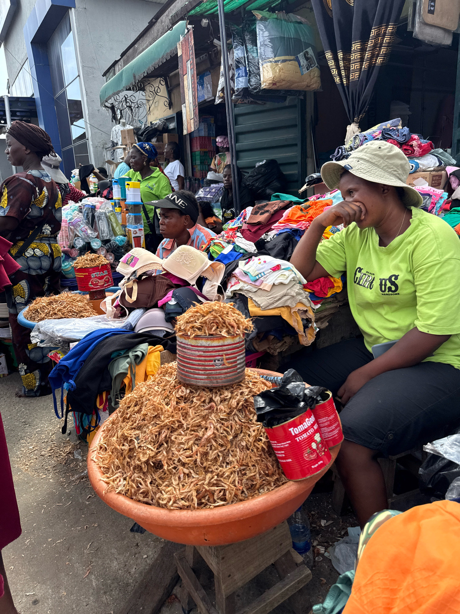 An outdoor market scene is captured in Lekki, Nigeria, with a focus on a vendor and her merchandise. In the foreground, a woman wearing a lime green t-shirt and a light-colored bucket hat sits on a stool. Her right hand is raised to her mouth, and she is looking away from the camera. Her t-shirt has the text "BEAUTIFUL US HANDSOME" printed on it. Next to her, on the left, is a large, orange basin overflowing with dried shrimp. Two red metal cans, one labeled "TomaGood TOMATO MIX", are placed on top of the shrimp.
Behind the woman, a variety of clothing and accessories are displayed. A stack of folded clothes, including colorful children's garments, is visible, along with bras and hats. Further back, other vendors and shoppers are present, with some of their faces partially visible. The market stall is covered by a dark fabric with a gold geometric pattern, and various items are hung from the stall's structure.
The ground is a rough, concrete surface scattered with debris. To the left of the frame, a woman in a brown and yellow patterned dress is partially visible, facing left. The overall impression is a busy, bustling marketplace.