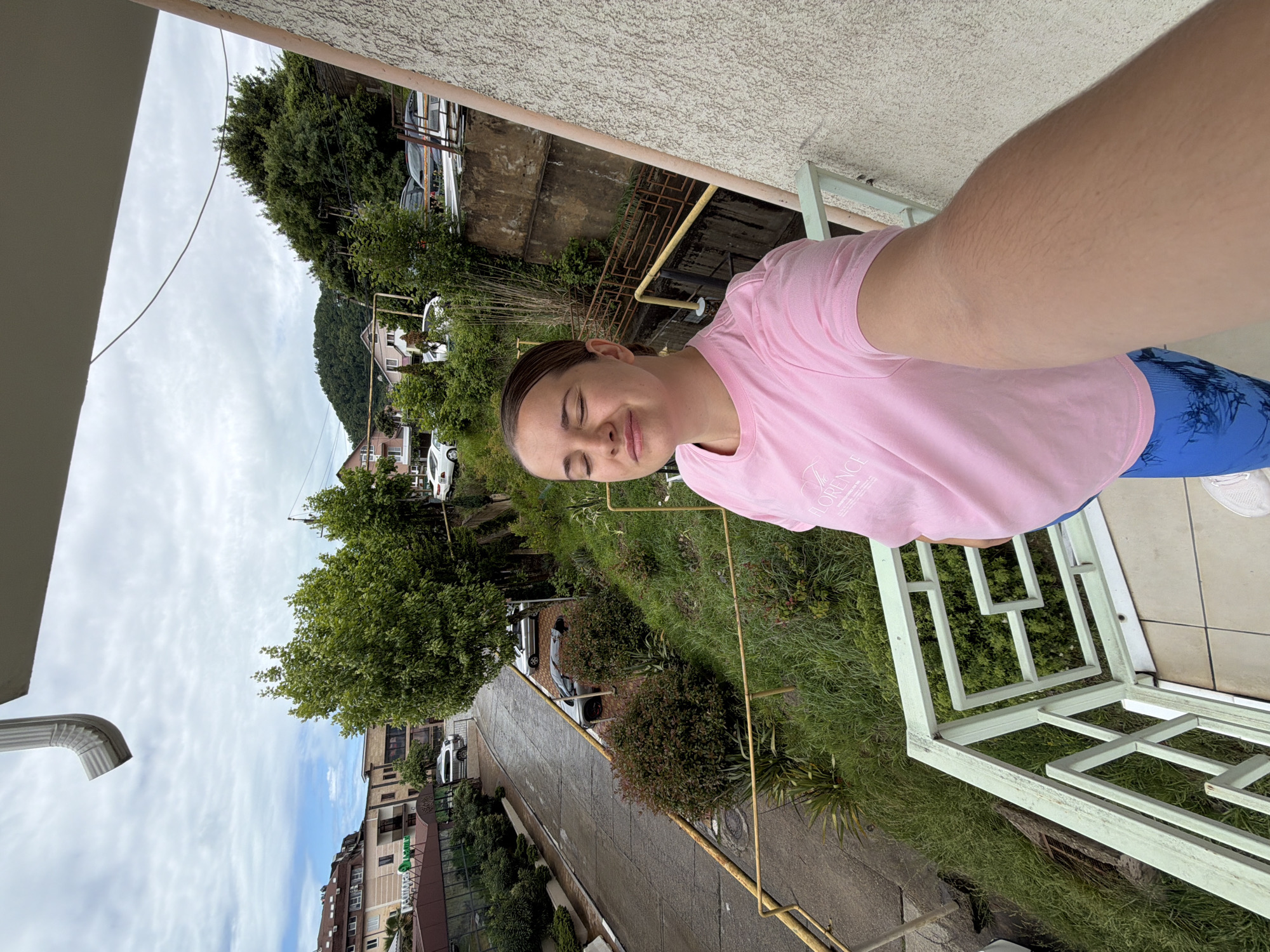 A woman takes a selfie on a balcony in Sochi, Russia. The woman is wearing a pink t-shirt and blue leggings. She is standing on a tiled balcony with a green railing. Behind her is a street with cars parked along it, and beyond that are buildings and trees. A mountain is visible in the distance. The sky is cloudy.