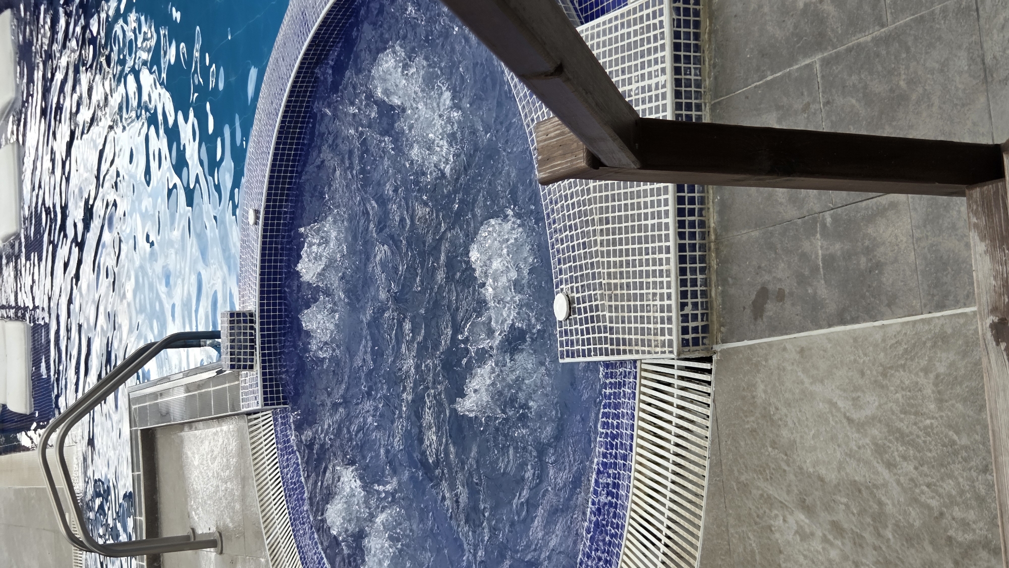 A high-angle, close-up shot captures a section of a blue tiled spa with bubbling water, adjacent to a larger blue swimming pool. Stainless steel handrails extend from the pool area into the spa. The spa's edge is lined with blue mosaic tiles, and a metal grate runs along the perimeter of the spa. The surrounding ground is covered in large, grey tiles. A wooden railing is visible in the foreground, to the right. The scene appears to be outdoors, possibly at a hotel or resort.