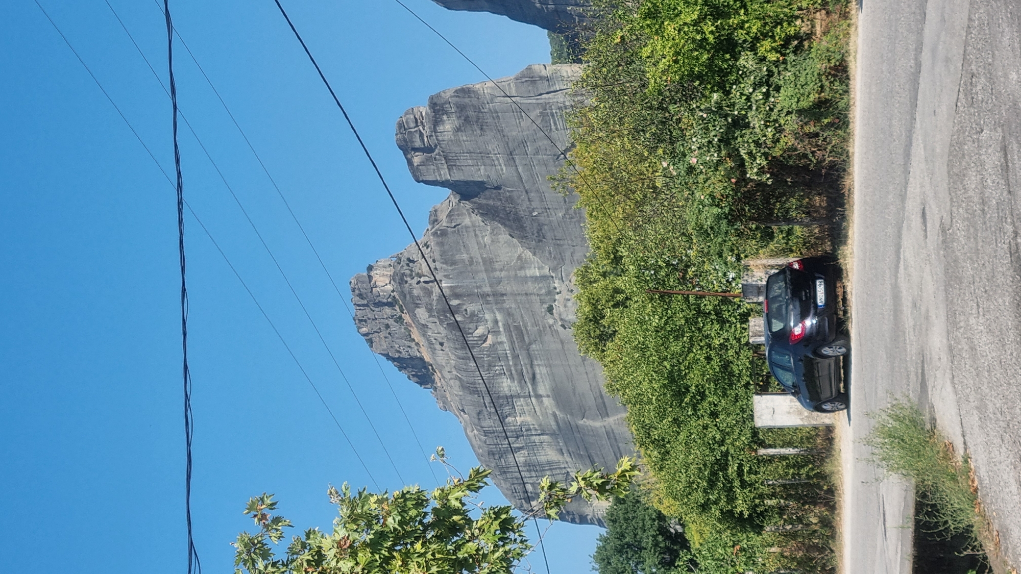 A black hatchback is parked on the side of a paved road in Kalambaka, Greece. The car is positioned perpendicular to the road, facing left. A rough-hewn stone fence runs behind the car, separating it from a dense green hedge. In the background, a massive, sheer rock formation with layered striations dominates the sky. Overhead, several dark electrical wires are strung across the clear blue sky. A leafy tree branch extends from the left side of the frame, partially obscuring the rock face.