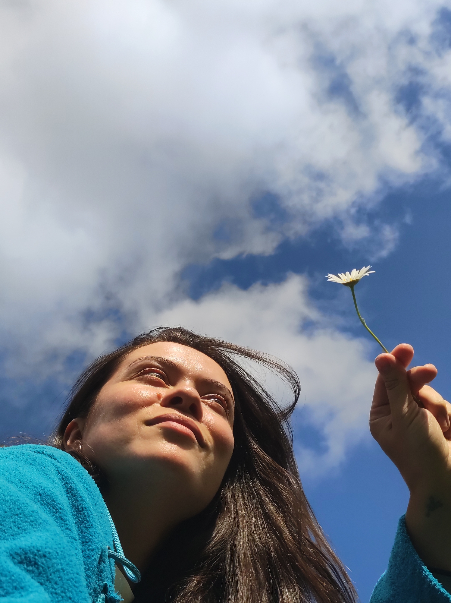 A low-angle shot shows a young woman looking up towards the sky, holding a single white daisy with a green stem. She is wearing a bright blue hooded garment. Her face is partially illuminated by sunlight. The background consists of a bright blue sky with scattered white clouds. There is no indication of the location being in the city of Фоки, Russia.